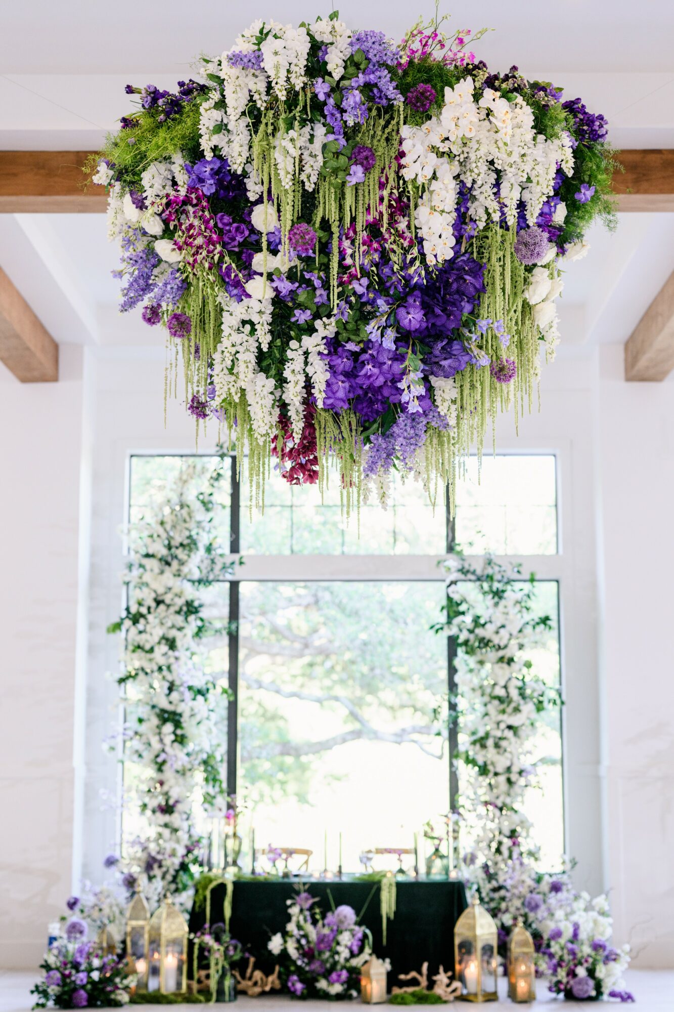 Hanging floral arrangement with white and purple flowers above a window with greenery and flowers below, in a bright room.