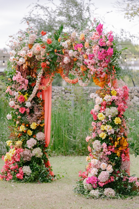Flower arch decorated with pink, white, and yellow flowers outdoors, with trees and sky in background.