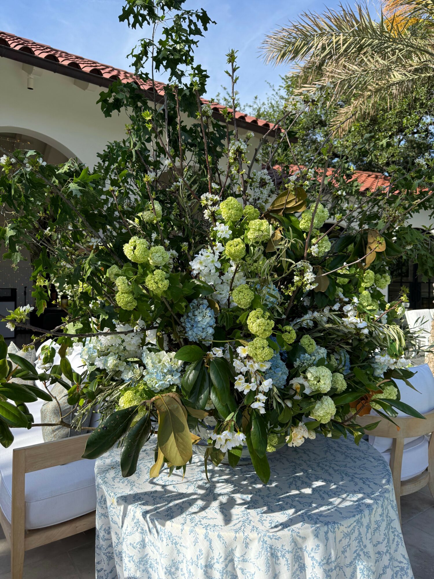 Large floral arrangement with white and green flowers on a table outdoors, with trees and a building in the background.