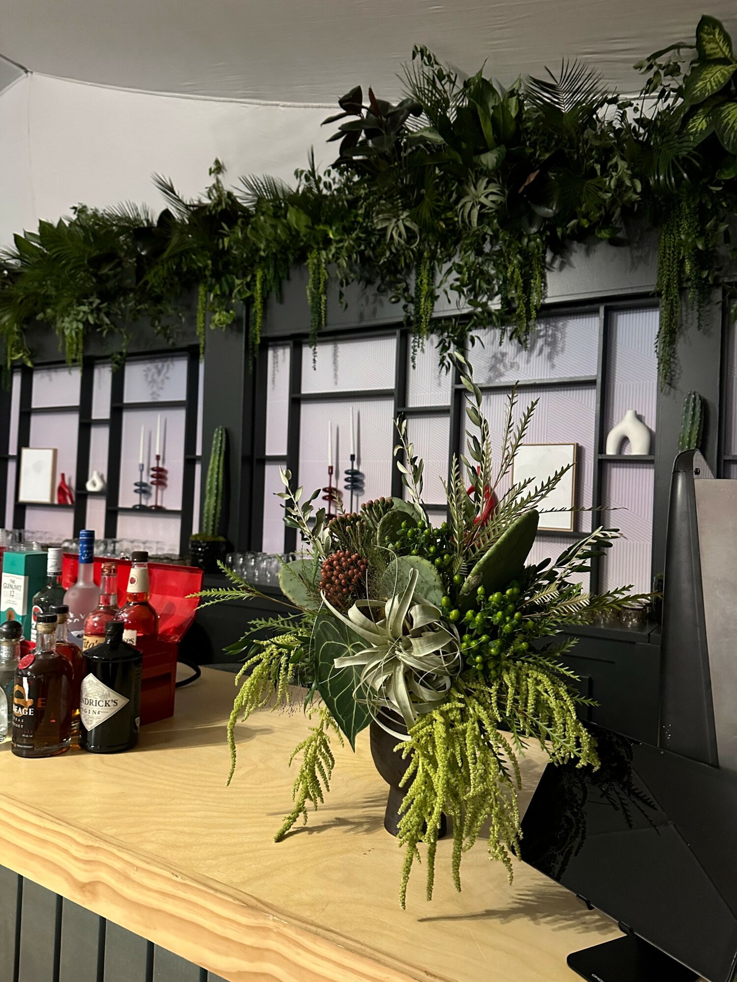 Flower arrangement with green foliage and succulents on a wooden table, with a decorated background and bottles nearby.