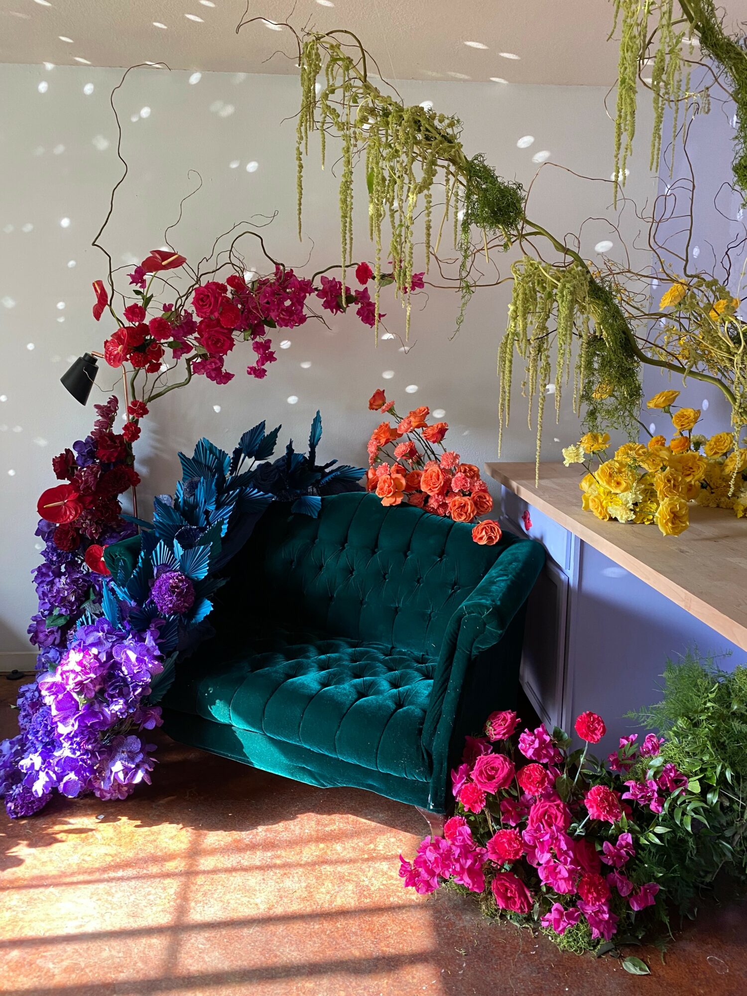 Teal velvet armchair surrounded by colorful flowers and hanging greenery, sunlight on the floor, floral display on a table.