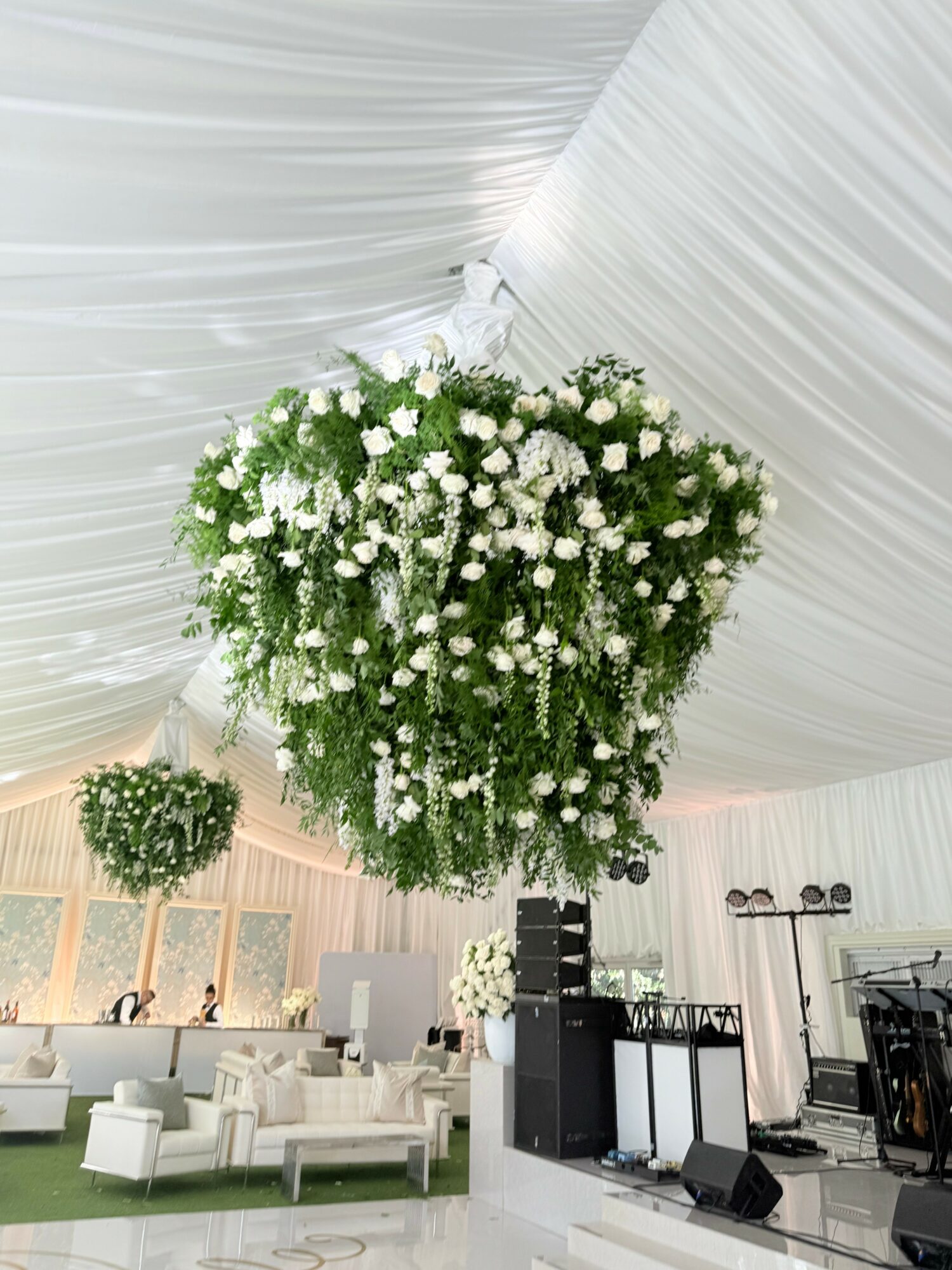 Large hanging floral arrangement with white flowers inside a decorated event tent, with seating and stage area visible.