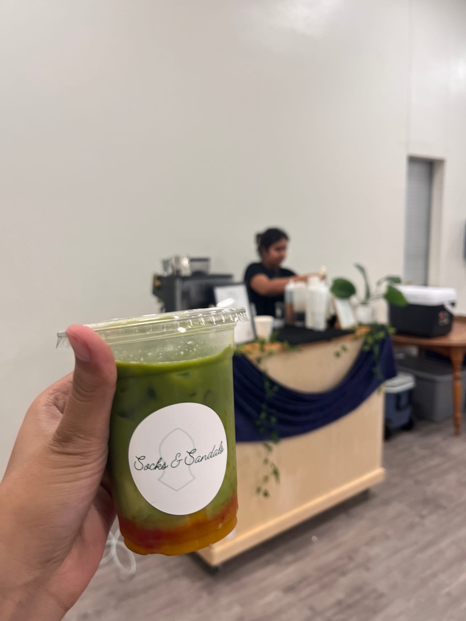 Hand holding a layered green and amber drink in a clear cup with a white label, background shows a person preparing beverages.