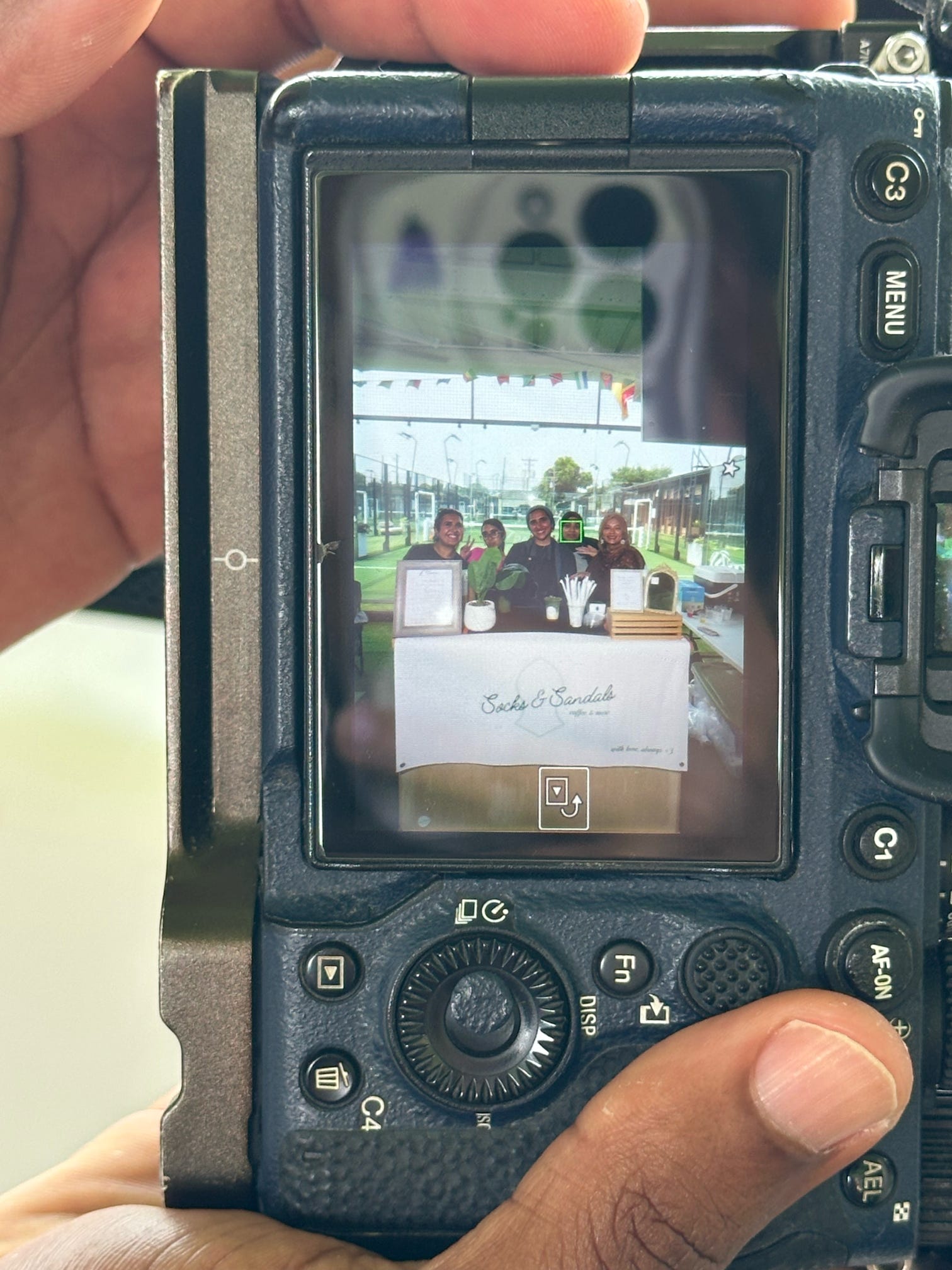 Person holding camera capturing a table with people outdoors, with a white sign on the table, background of trees and sky.