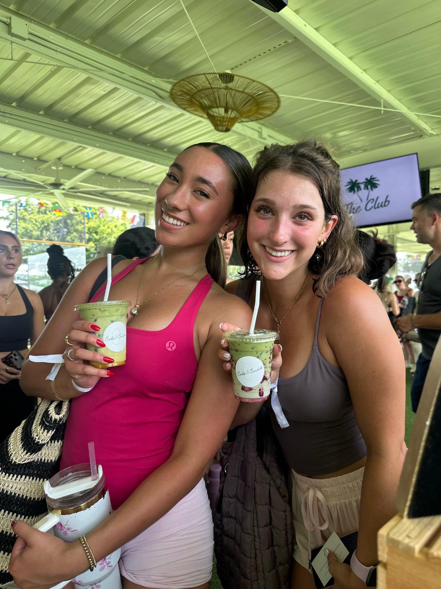 Two women smiling and holding drinks in a lively outdoor setting with a thatched umbrella and a TV screen in the background.