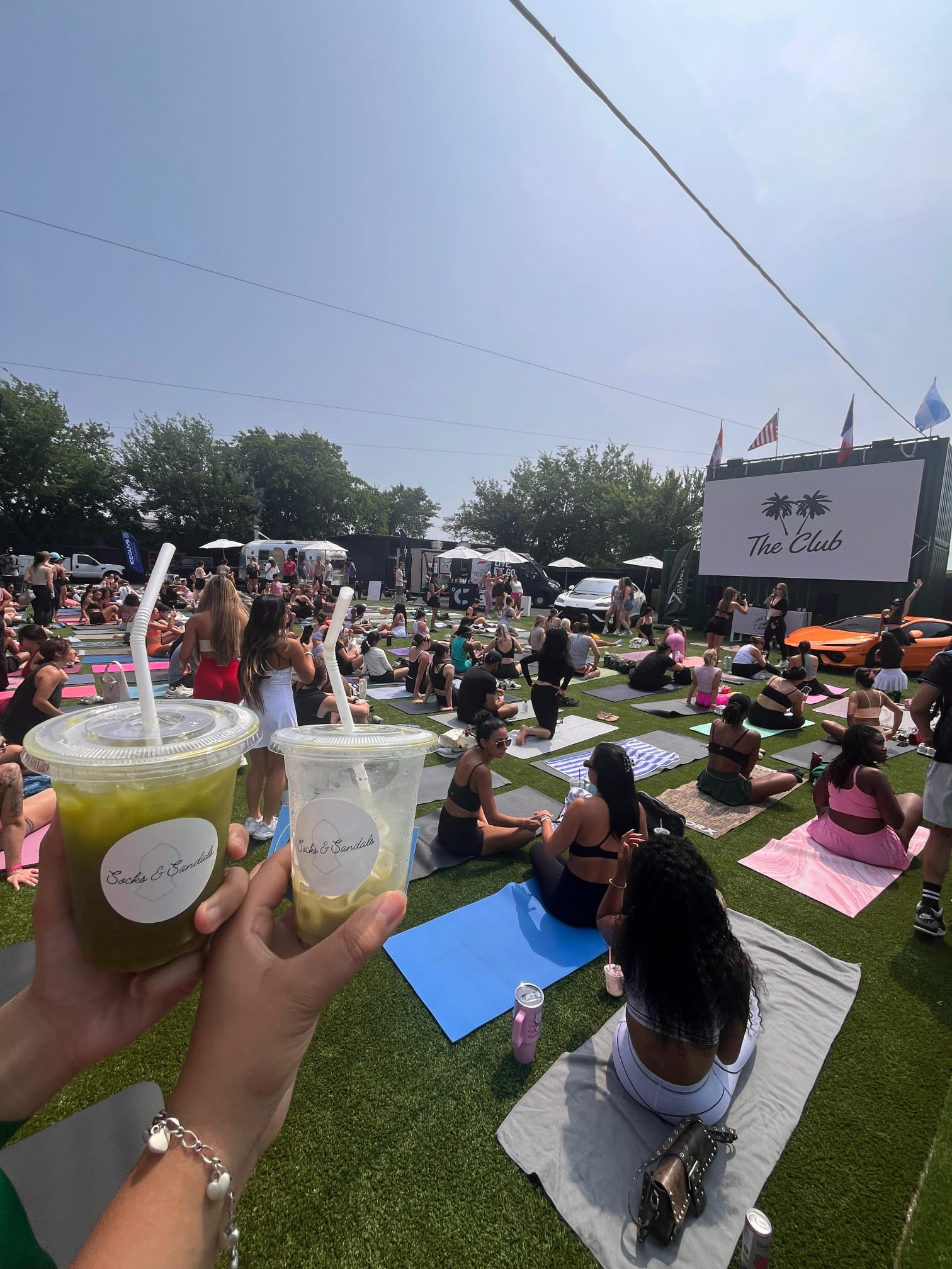 People sitting on grass with yoga mats watching a large outdoor screen, holding drinks, sunny day, trees in background.