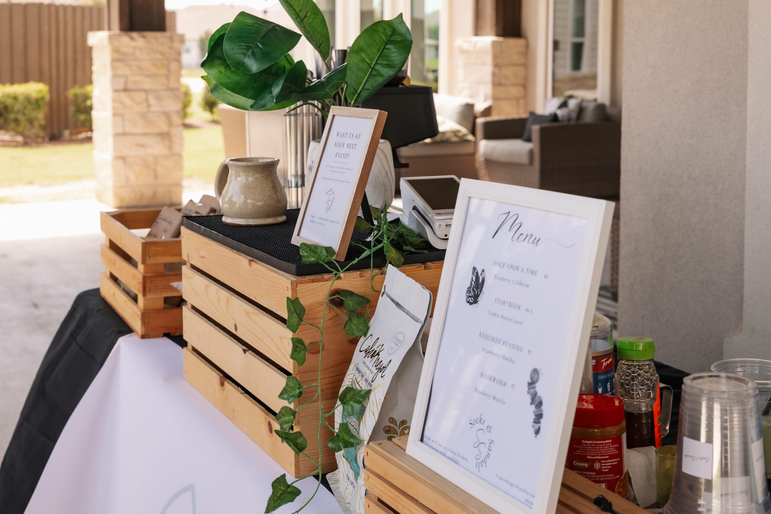 Table with framed menu, potted plant, and condiments, outdoors with a building and porch in background.