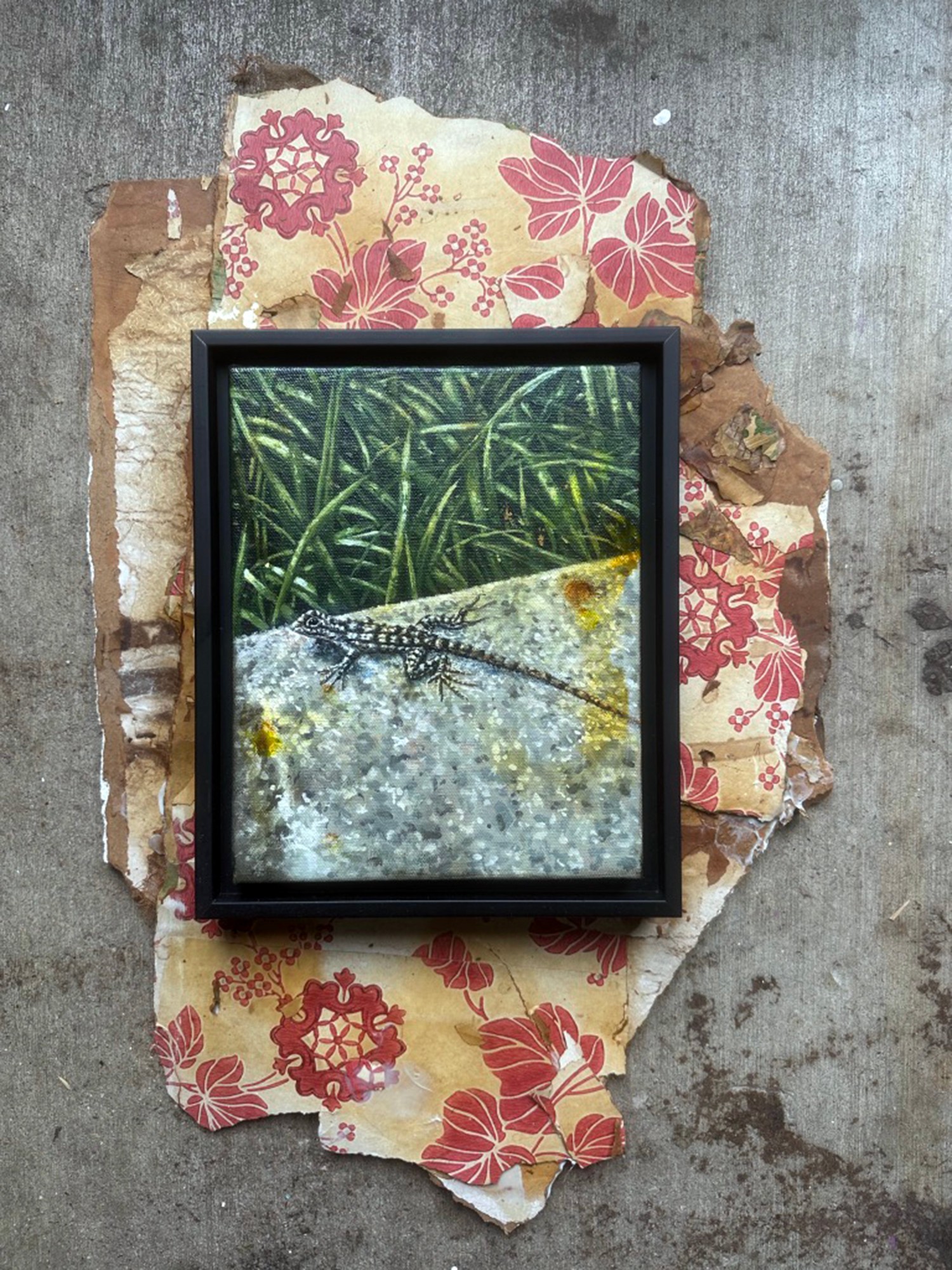 Framed picture of a crocodile on grass, placed on torn floral paper and concrete surface.