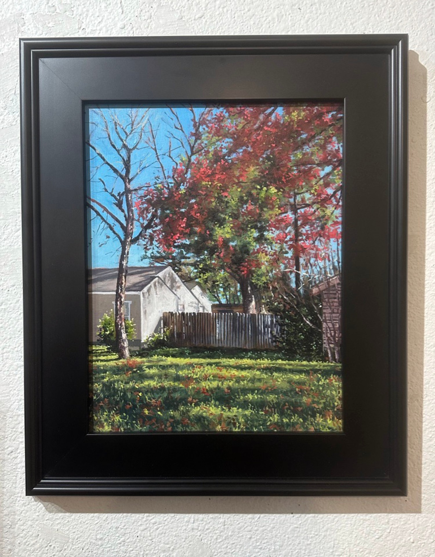 Framed landscape photo of a backyard with trees, a fence, and a house, hanging on a white wall.