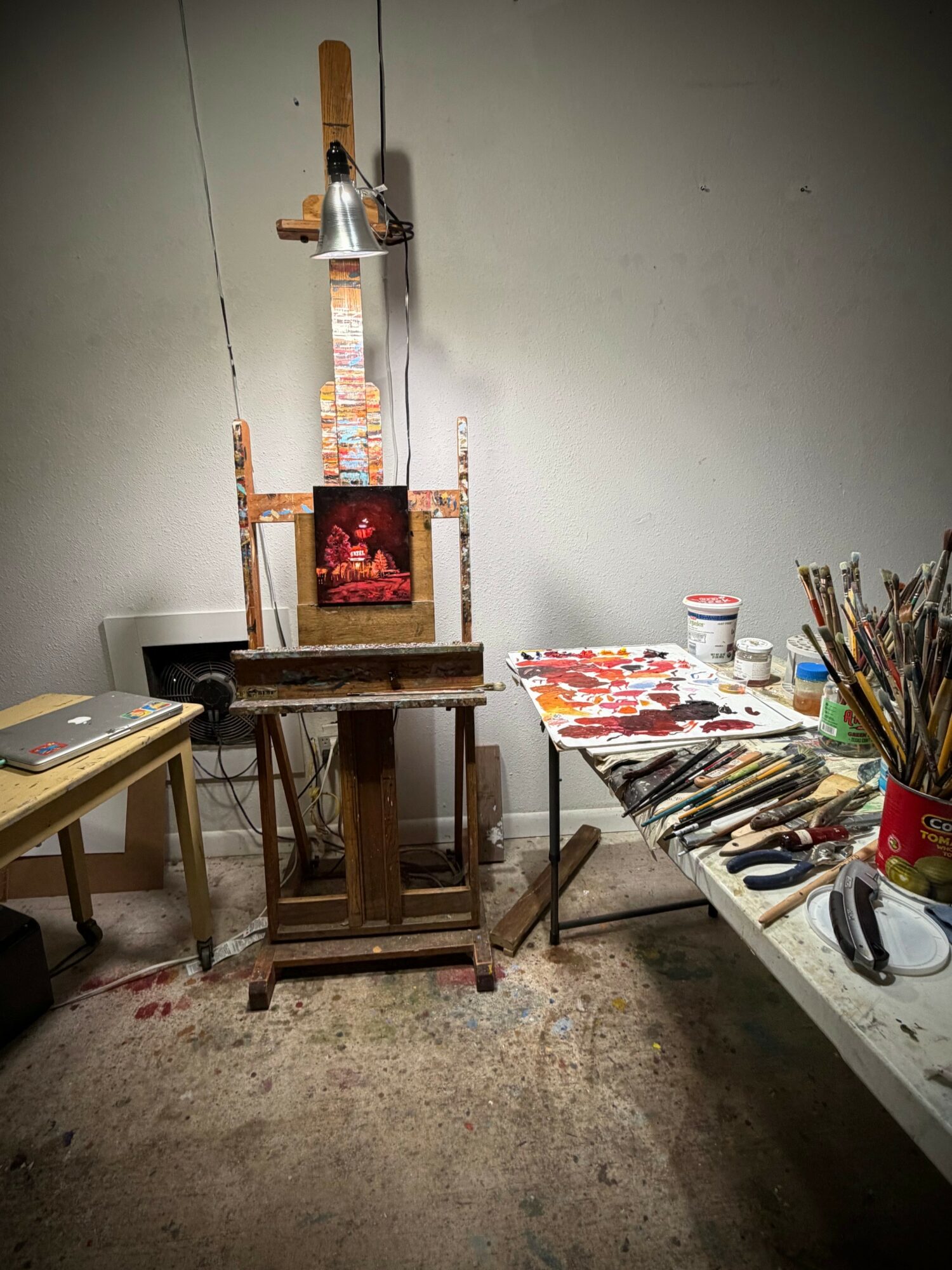 Art studio with easel, painting supplies, and a table with brushes and paints, against a plain wall.
