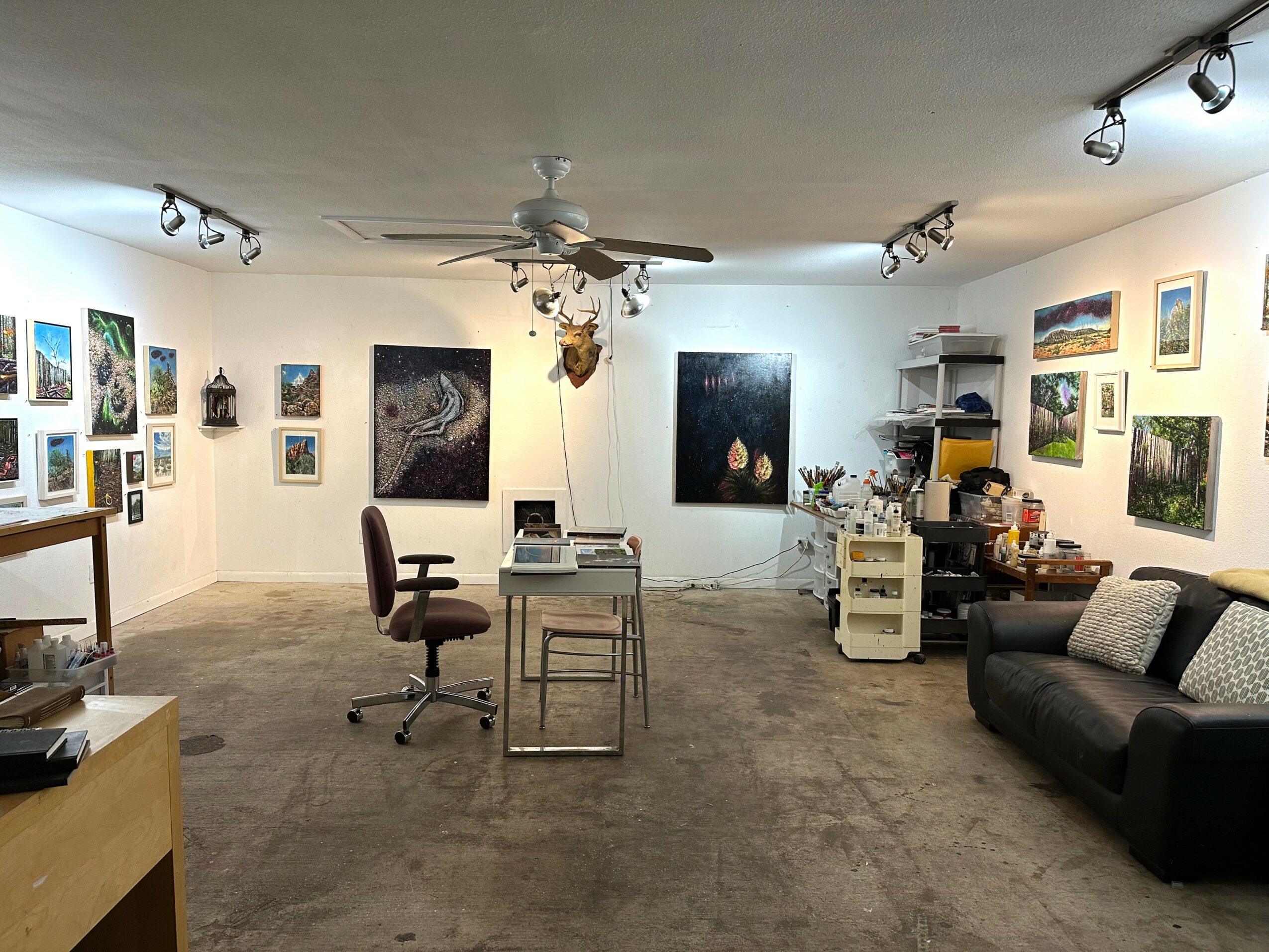 Gallery room with paintings on white walls, a black sofa, a desk with a chair, and a ceiling fan.