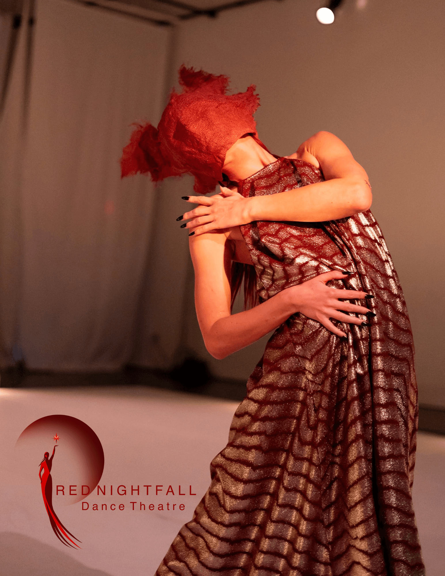Performer in a patterned dress and red headpiece dances on stage with arms crossed, background with curtains and stage lighting.