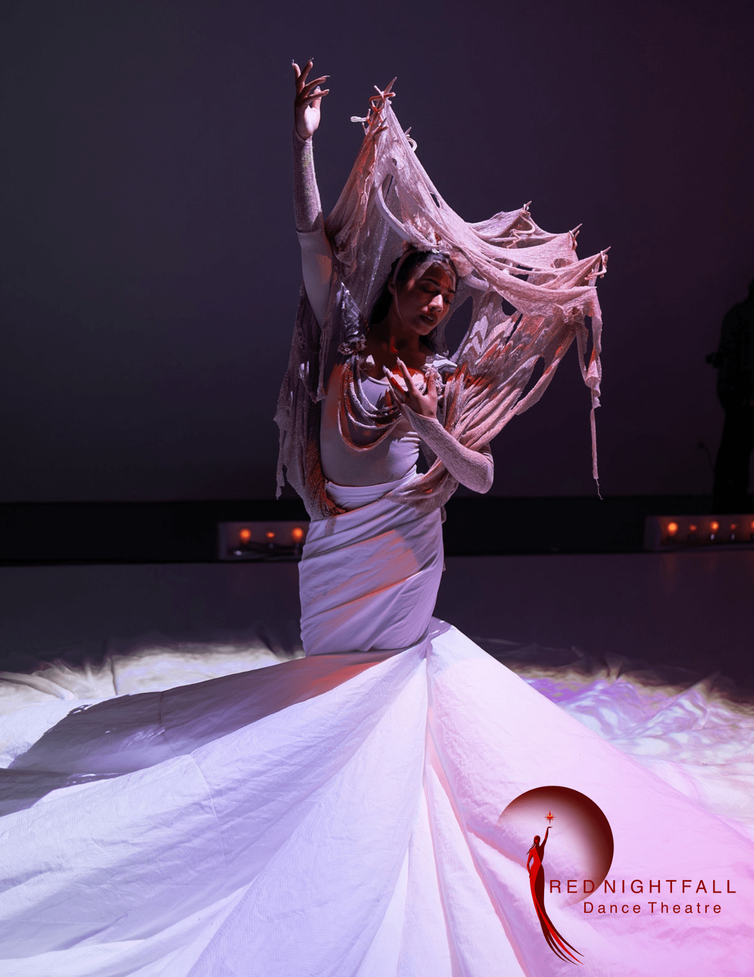 Performer in a flowing white costume with fabric extending outward, raising one arm, on stage with dark background.