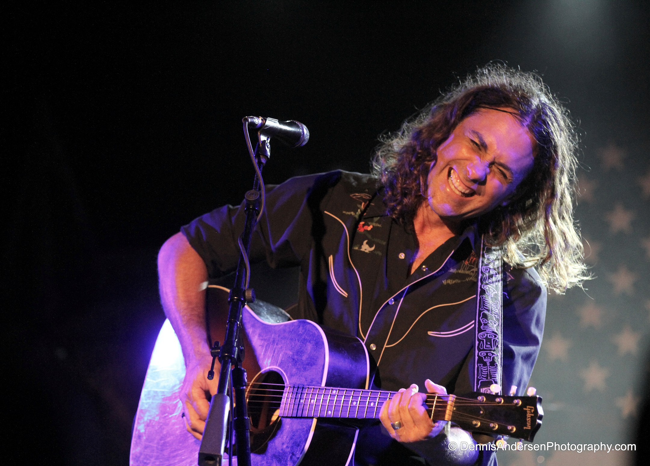 Person with curly hair playing an acoustic guitar on stage, smiling, with a microphone nearby.