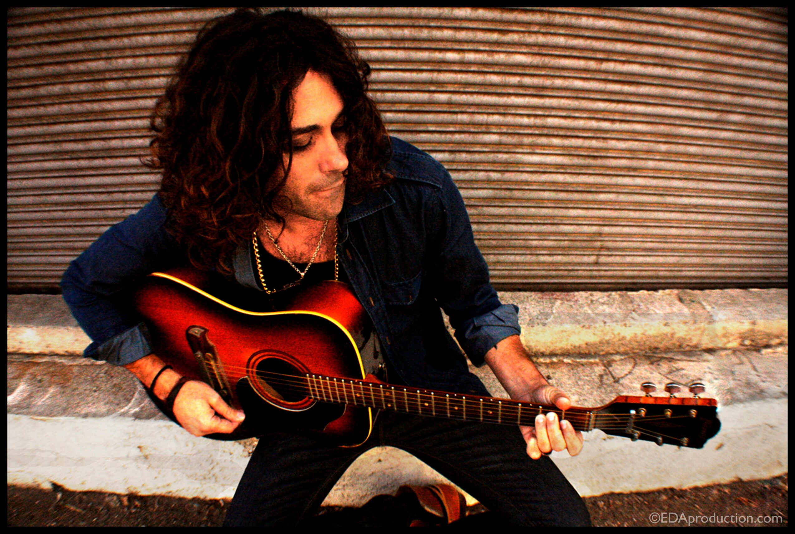 Person with curly hair playing an acoustic guitar outdoors against a textured background.