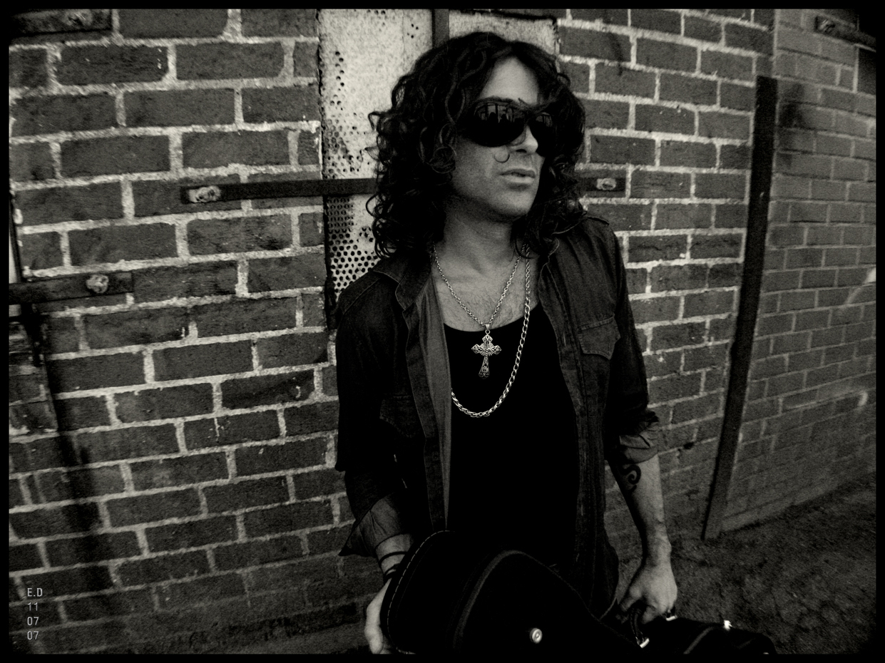 Woman with curly hair wearing sunglasses and a necklace, standing against a brick wall.