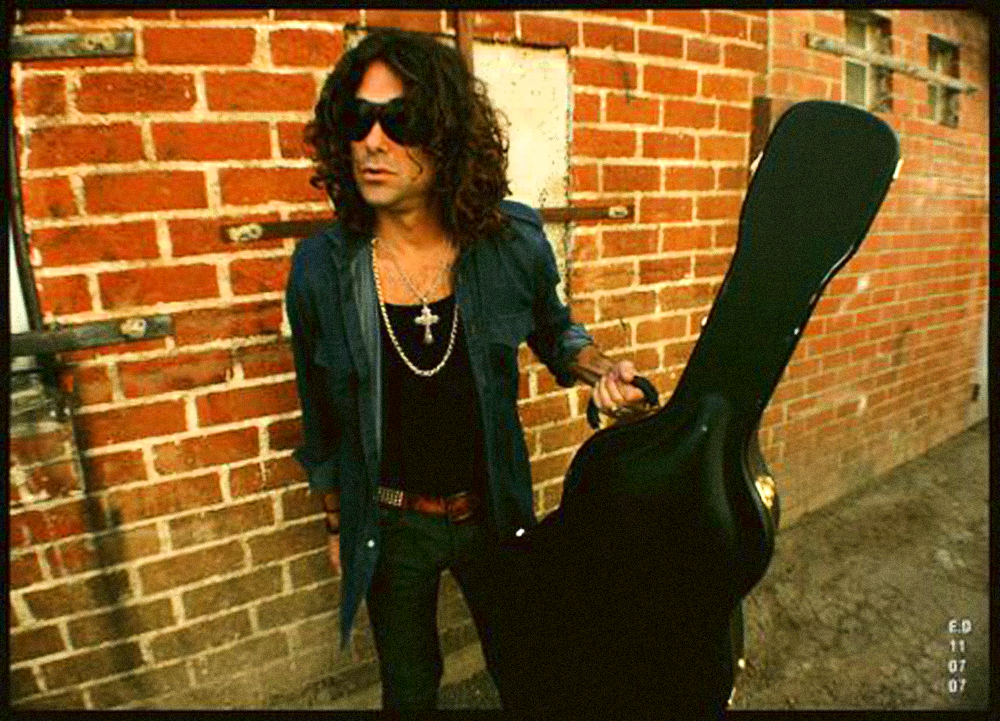 Person with curly hair, sunglasses, and dark clothing holding a guitar case against a brick wall.
