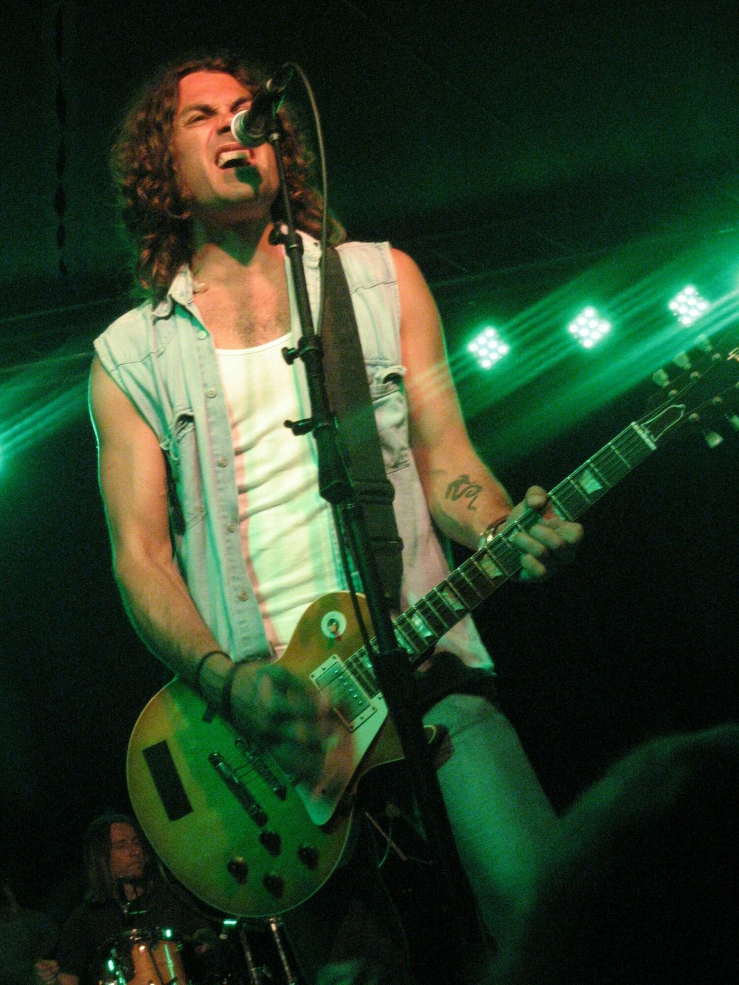 Person playing guitar on stage with green lighting, wearing sunglasses and a light-colored shirt, singing into a microphone.