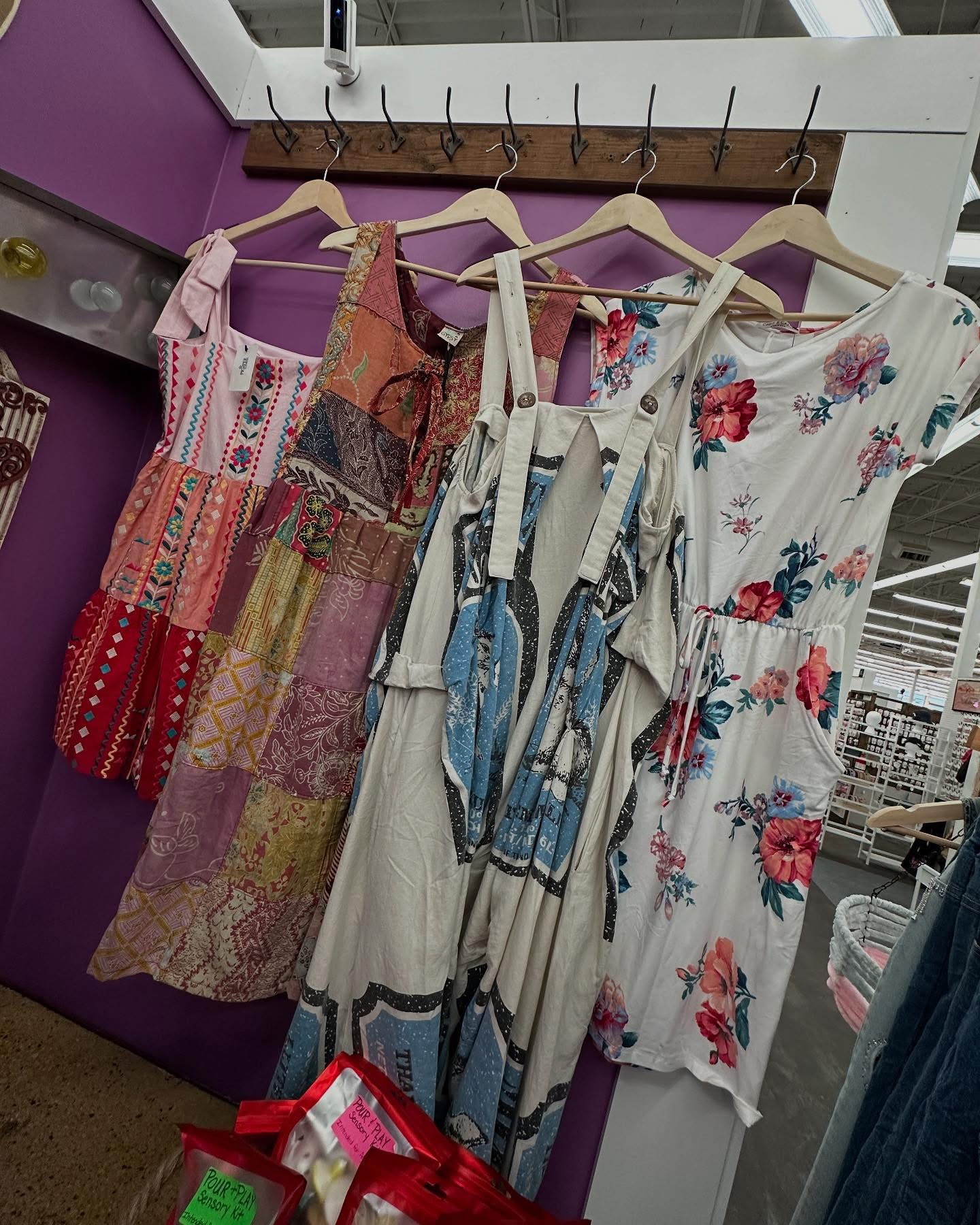 Four dresses hanging on a rack with floral and patterned designs, displayed indoors against a purple wall.