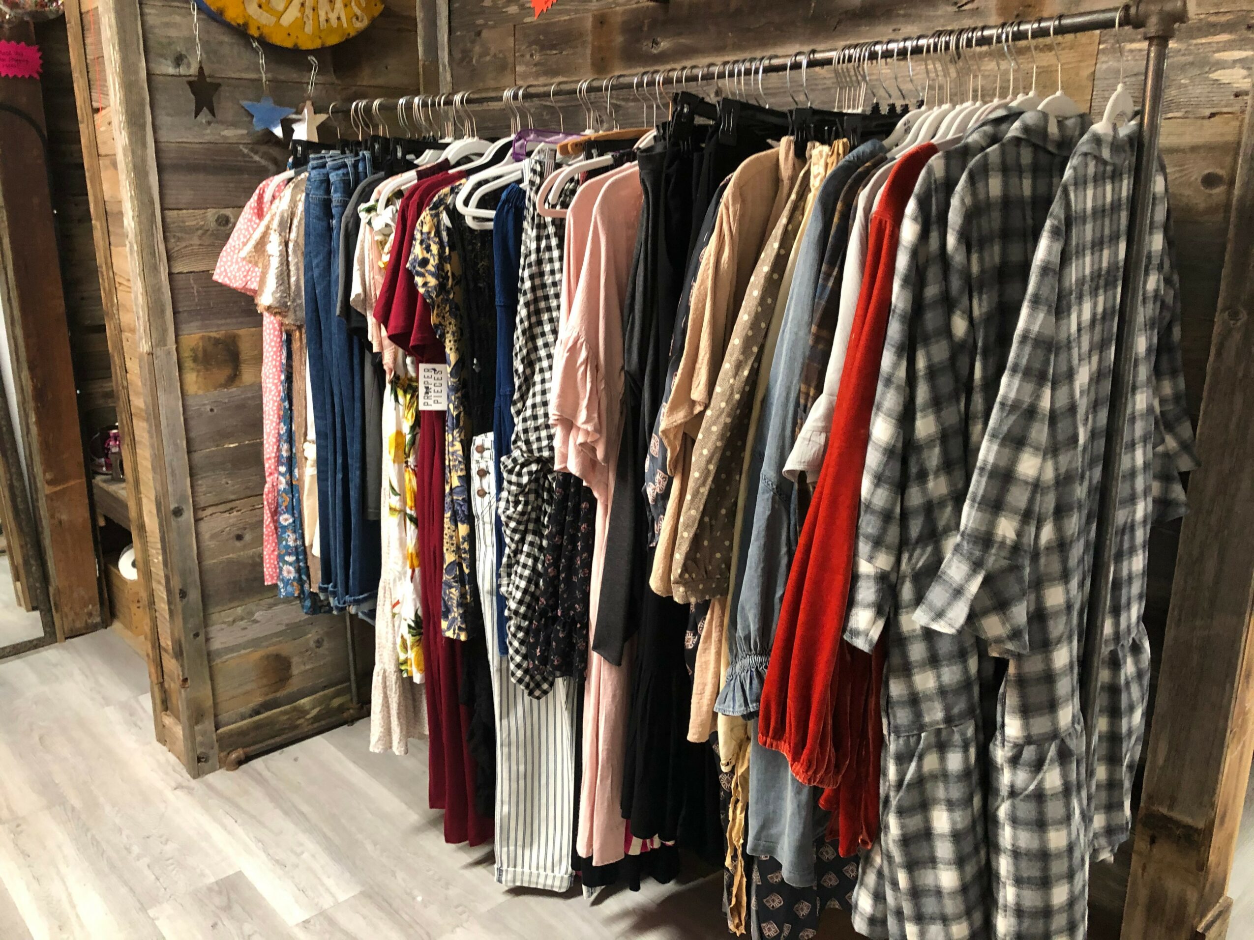 Clothing rack with various dresses, shirts, and garments hanging on a metal rod in a rustic store setting.