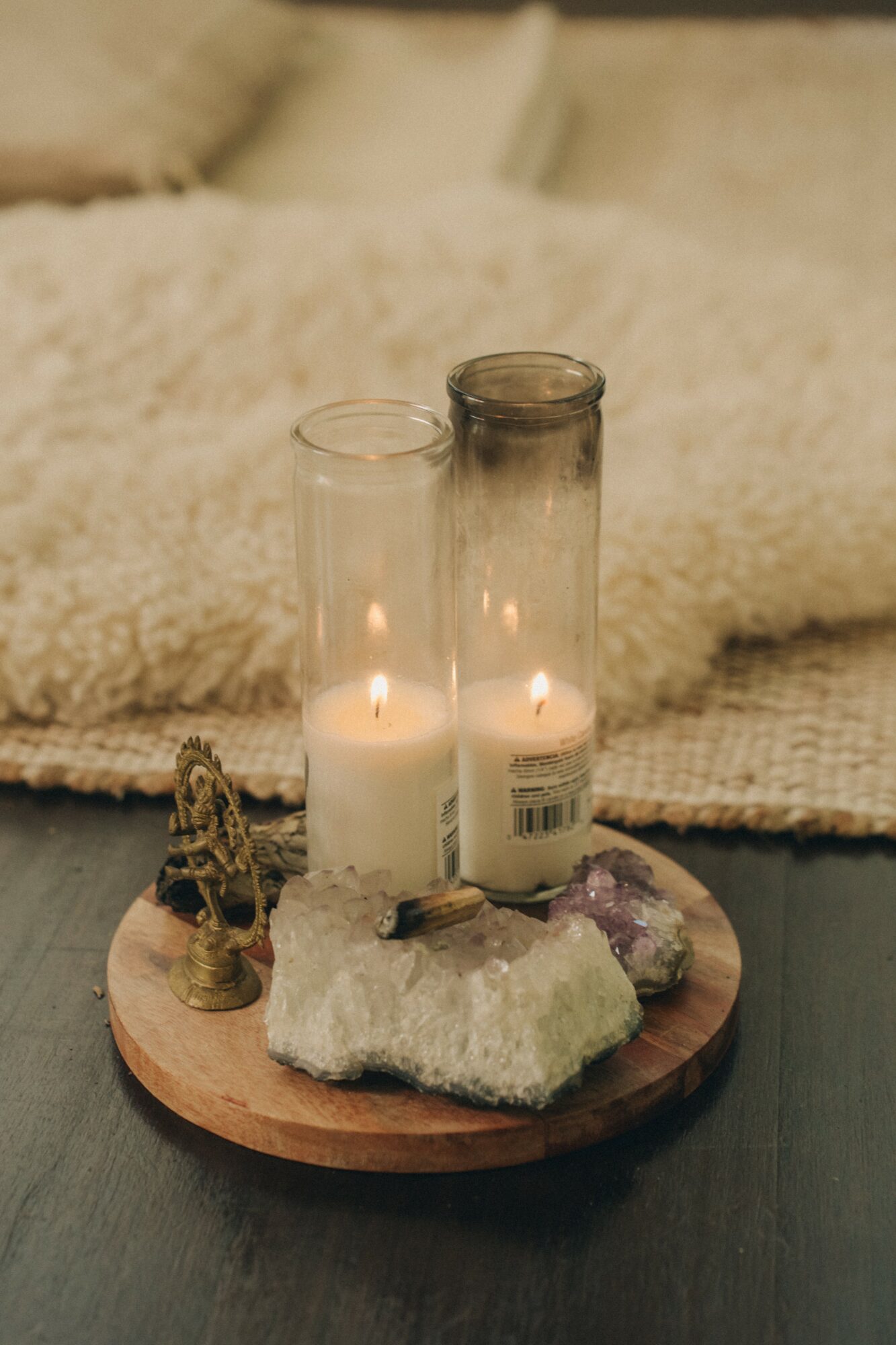 Two lit candles in glass holders on a wooden tray with crystals and a small figurine, against a soft background.