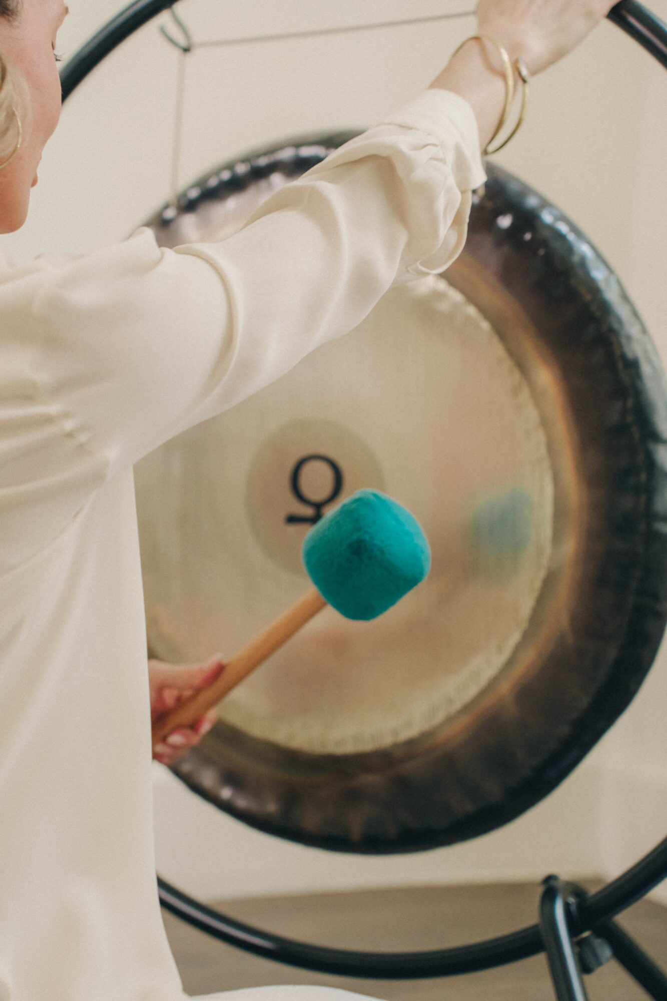Person playing a large gong with a mallet, wearing a light-colored shirt, in a music or performance setting.