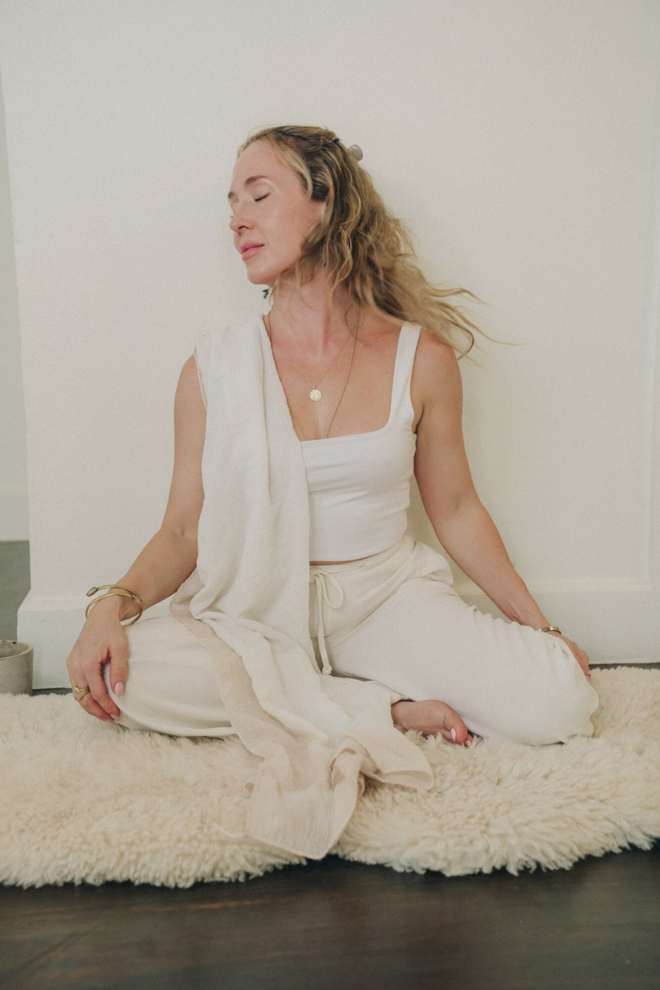 Woman sitting cross-legged on a rug with eyes closed, wearing white clothing, against a plain white wall.
