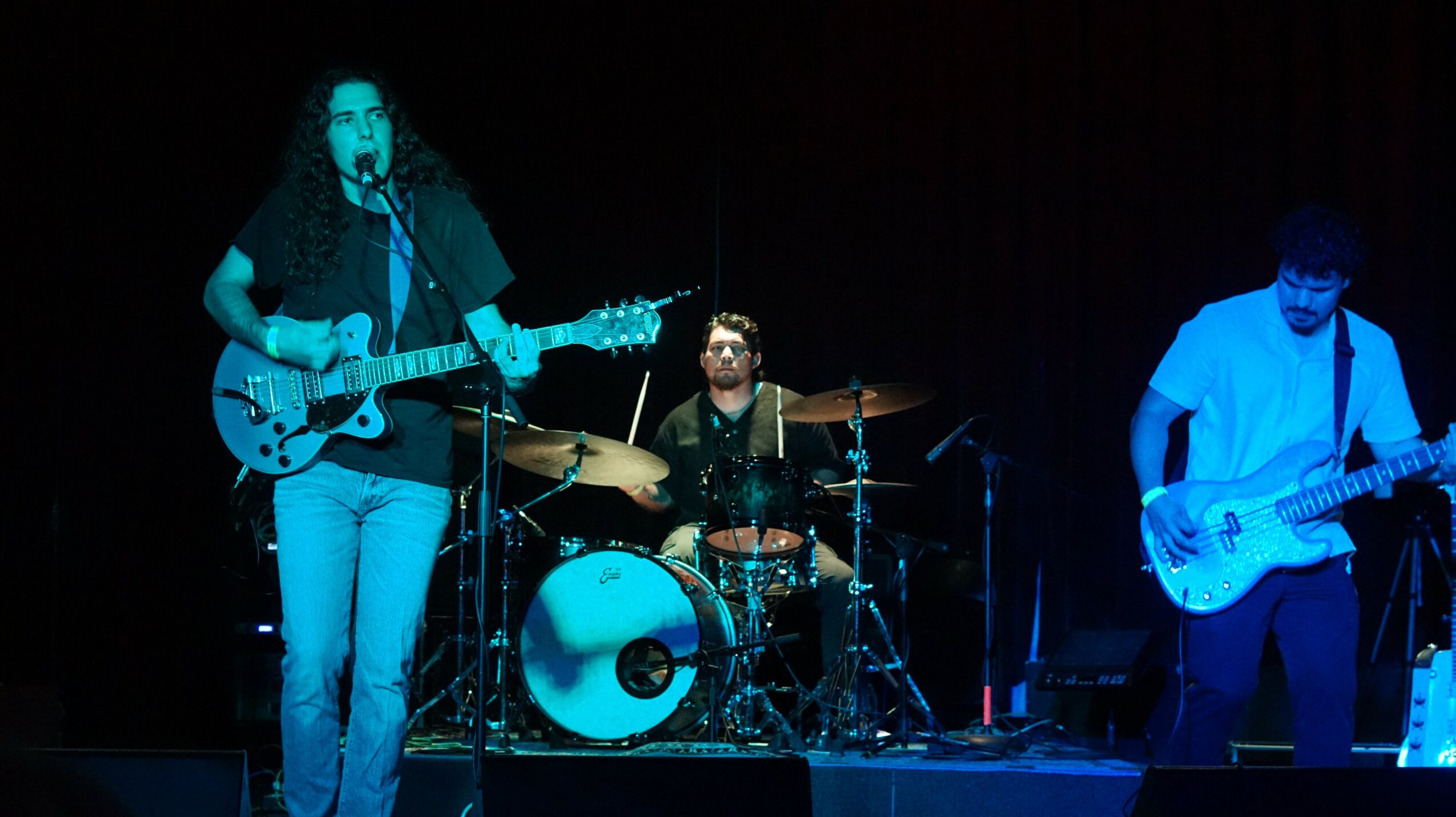 Three musicians perform on stage with a guitar, drums, and bass guitar, under dark lighting.
