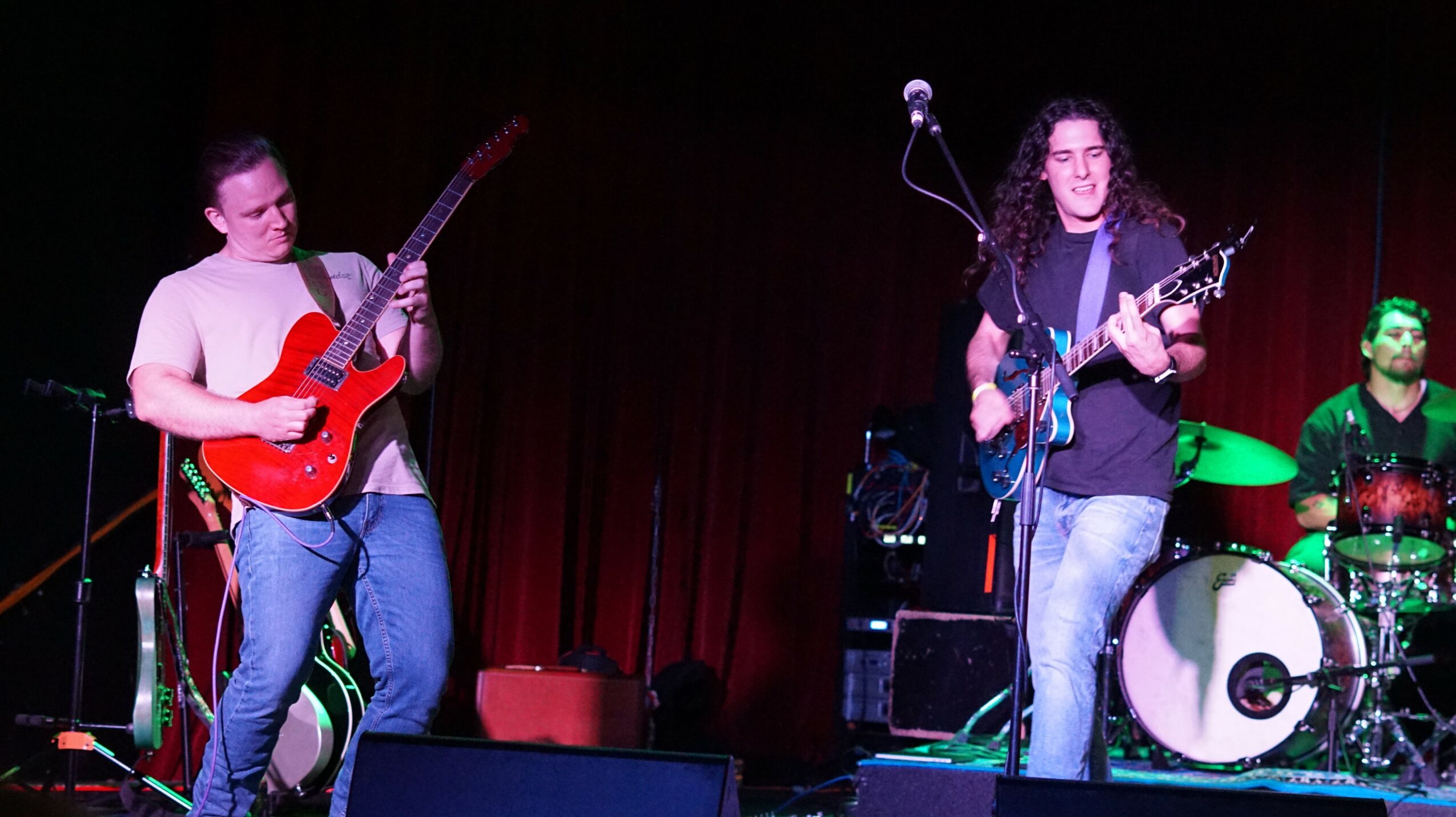 Three musicians perform on stage with red curtain background; two play guitars, one drums, with microphone and equipment visible.