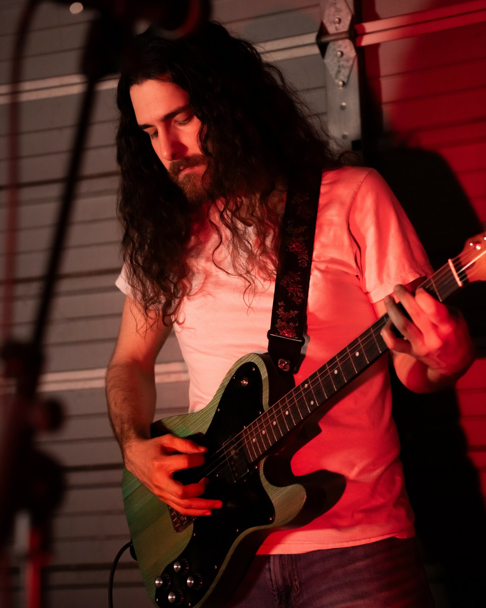 Person with long curly hair playing an electric guitar, wearing a light-colored shirt, in a dimly lit setting.