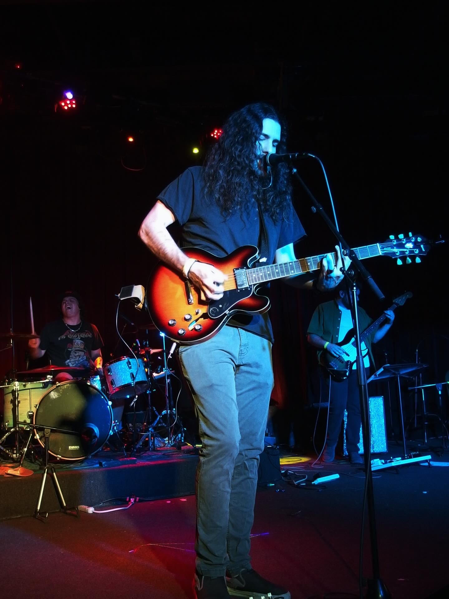 Musician with long curly hair playing an electric guitar on stage with band members, dark background, colorful stage lights.
