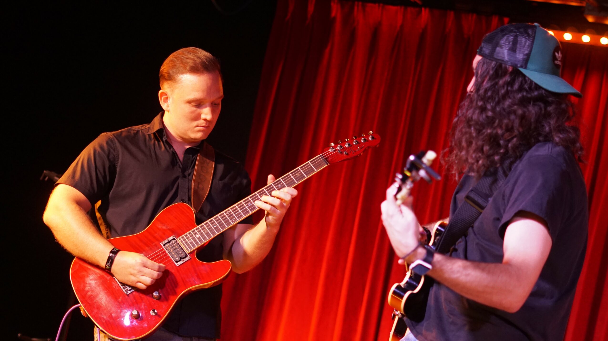 Two musicians playing guitars on stage with red curtains in the background.