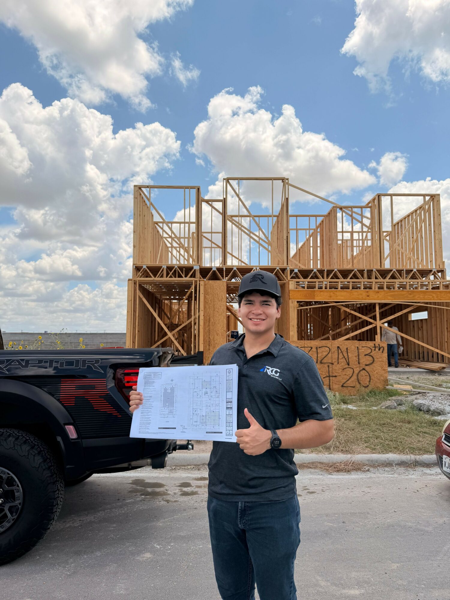 Construction worker holding a clipboard in front of a wooden building frame under a cloudy sky.
