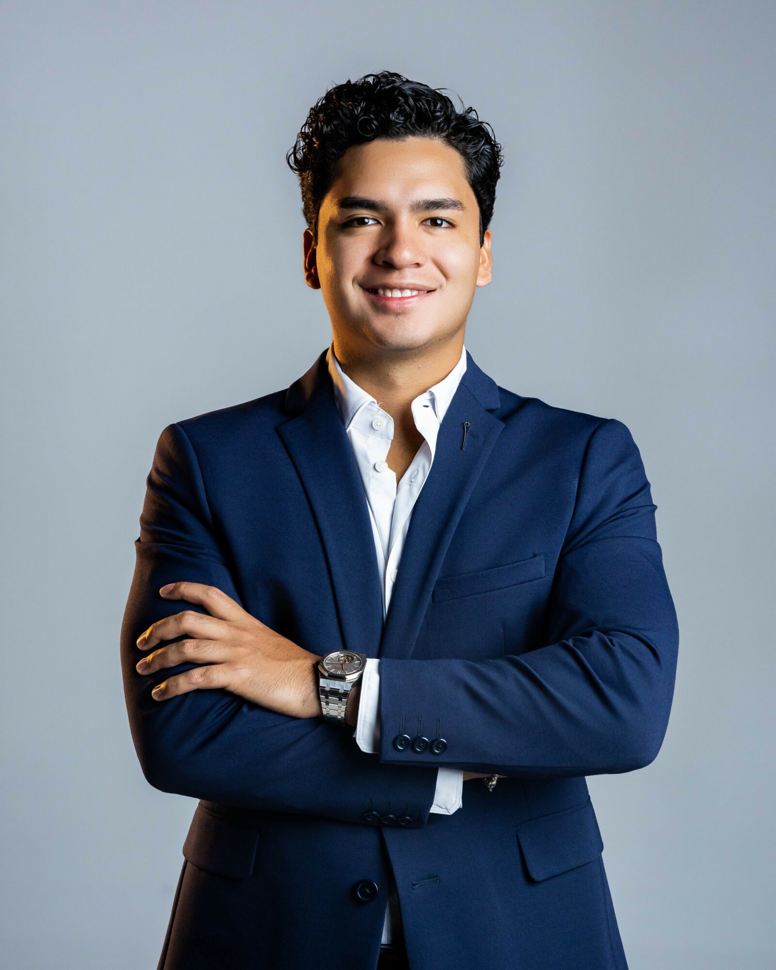 Young man in a navy suit with arms crossed, smiling, against a plain light gray background.