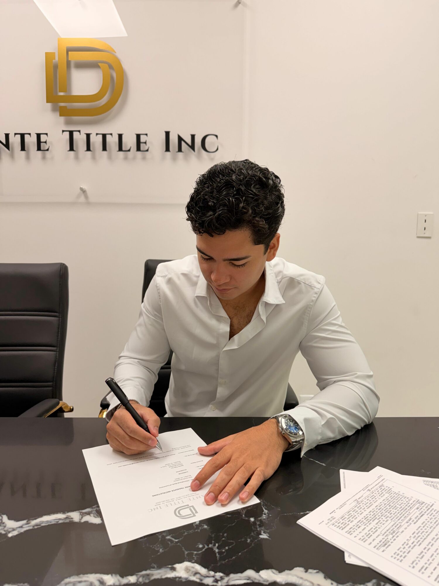 Man in white shirt writing on paper at desk with documents, in office setting, with company logo on wall