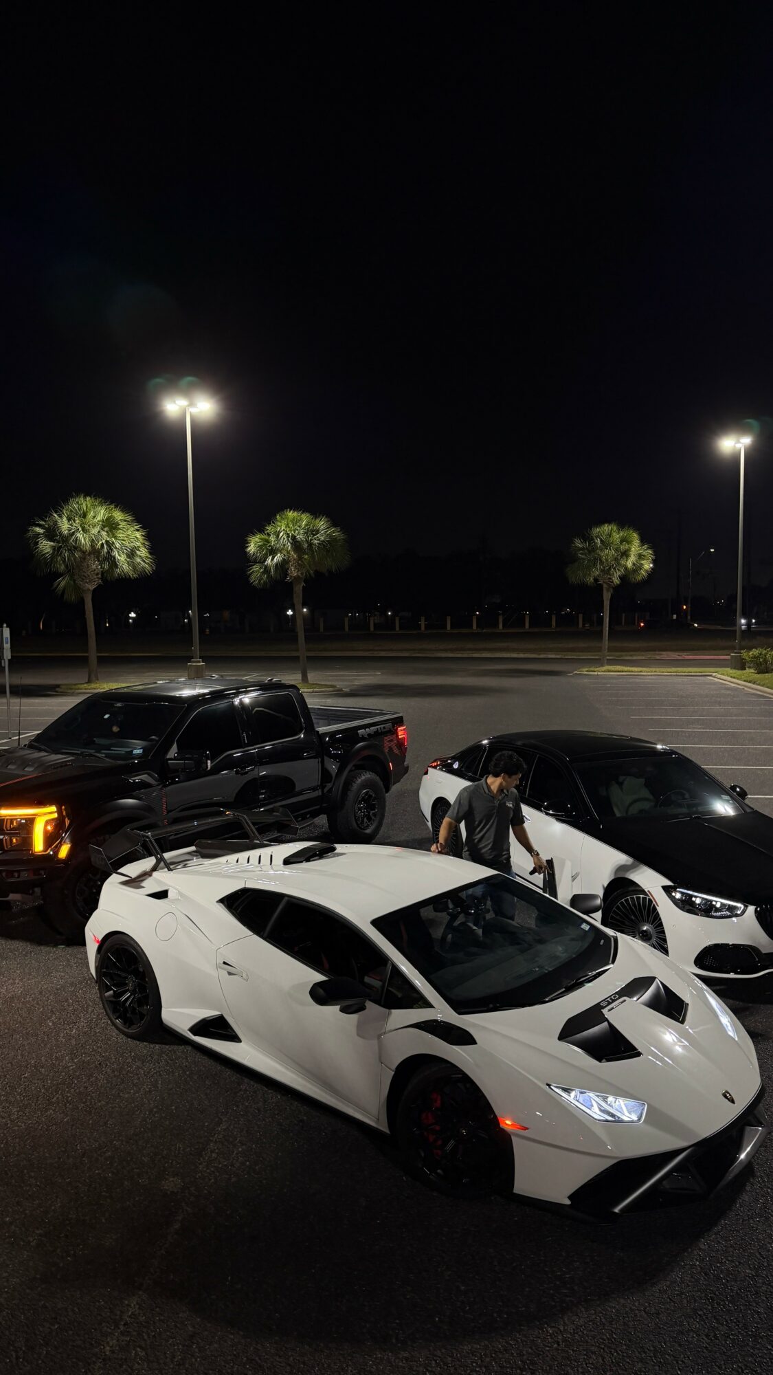 Night scene with three parked cars and two people near a white sports car, illuminated by streetlights, with trees in the background.