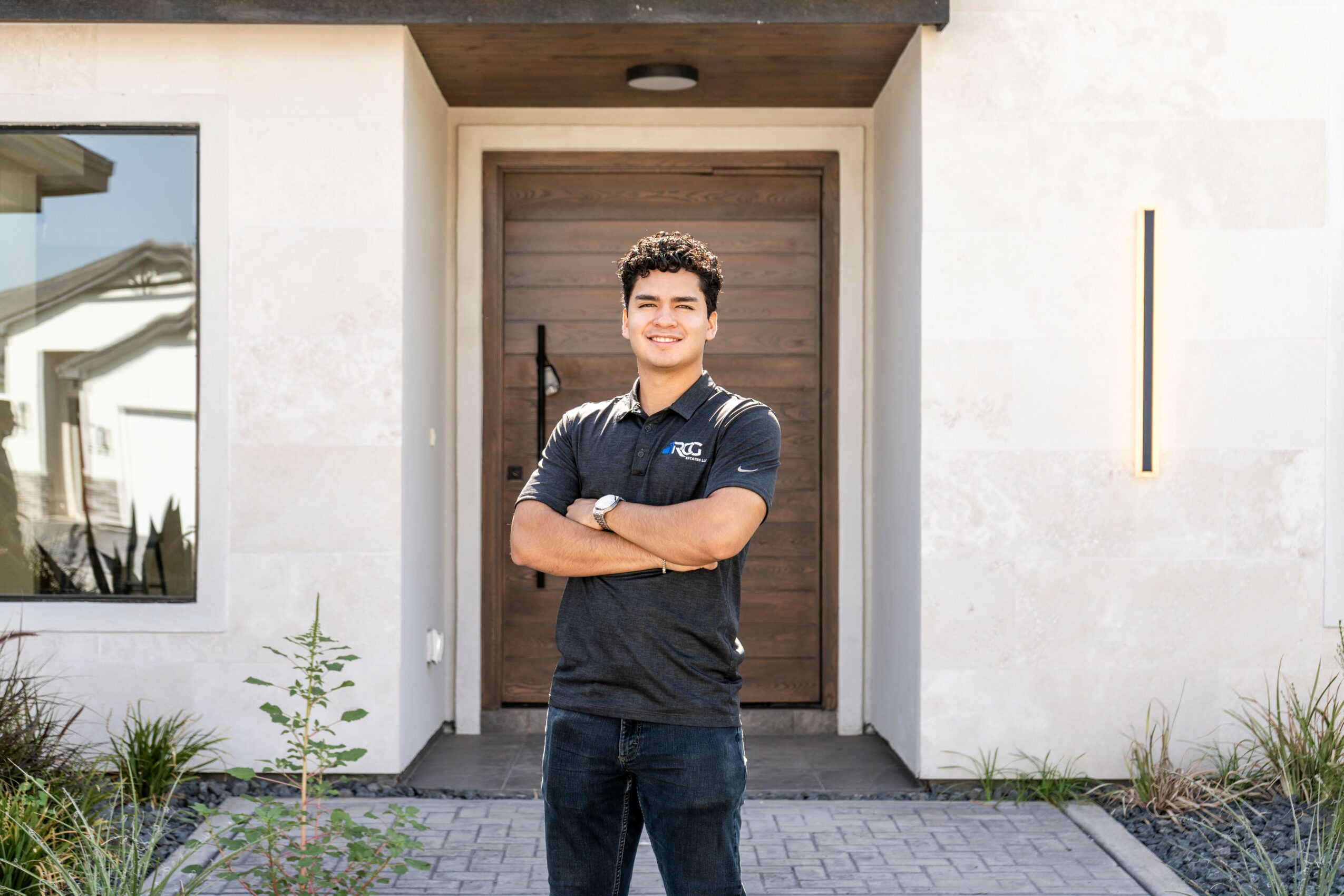 Young man standing with arms crossed in front of a modern house entrance, smiling, wearing a dark polo shirt and jeans.