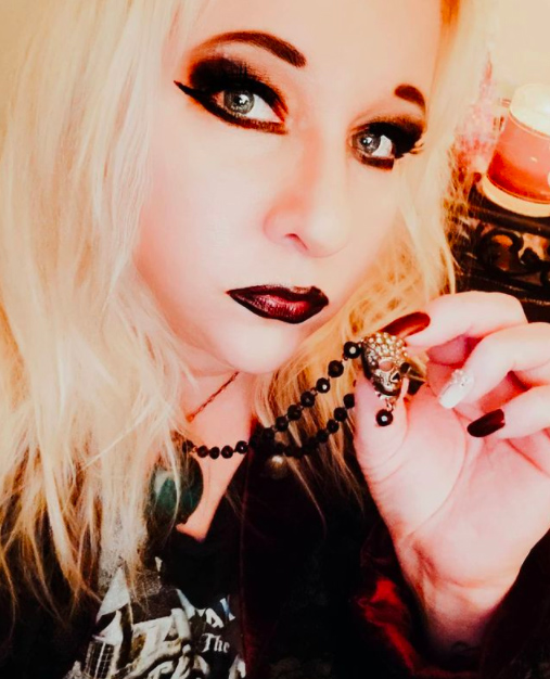 Close-up of a woman with platinum blonde hair, dark makeup, and black lipstick holding a beaded necklace, with a pink candle in the background.