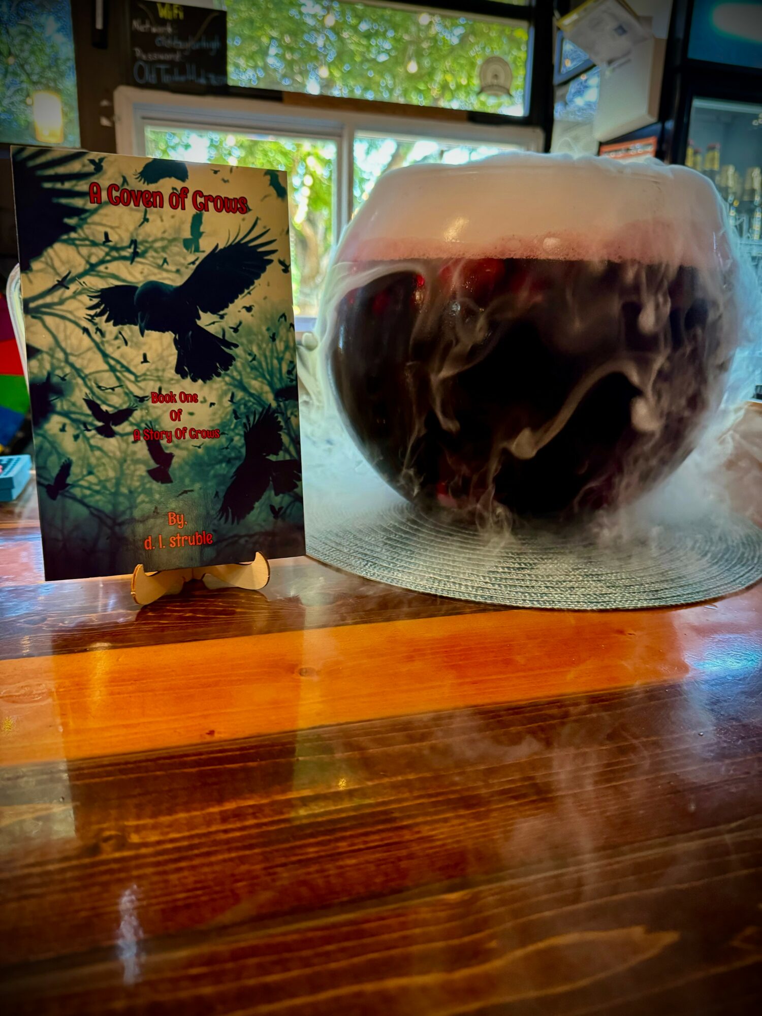 Book titled 'Crown of Crows' and a glass of dark beer with foam on a wooden table, with windows in the background.