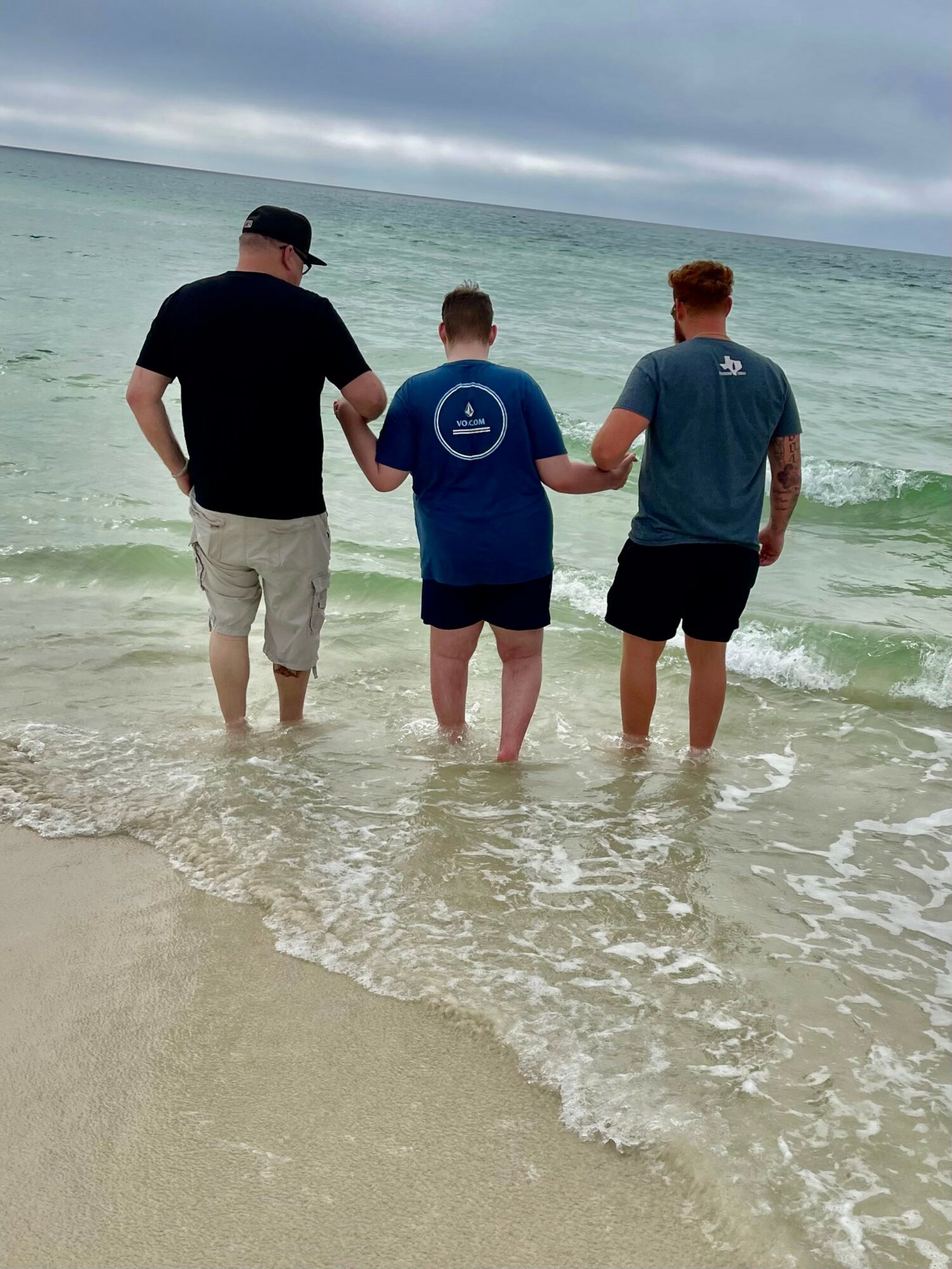 Three people holding hands and walking into the ocean on a cloudy day, with waves and sandy beach visible.