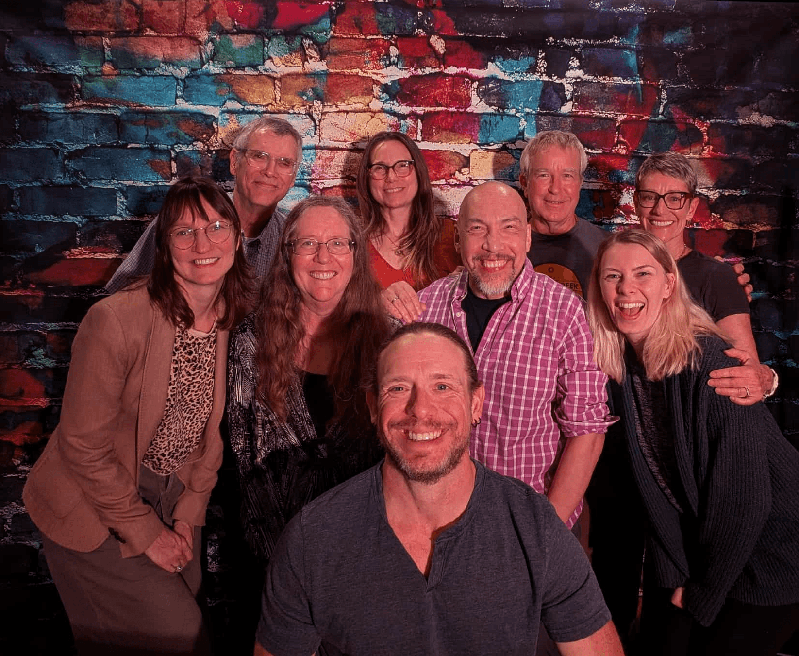 Group of ten people smiling in front of a colorful brick wall, some with arms around each other.