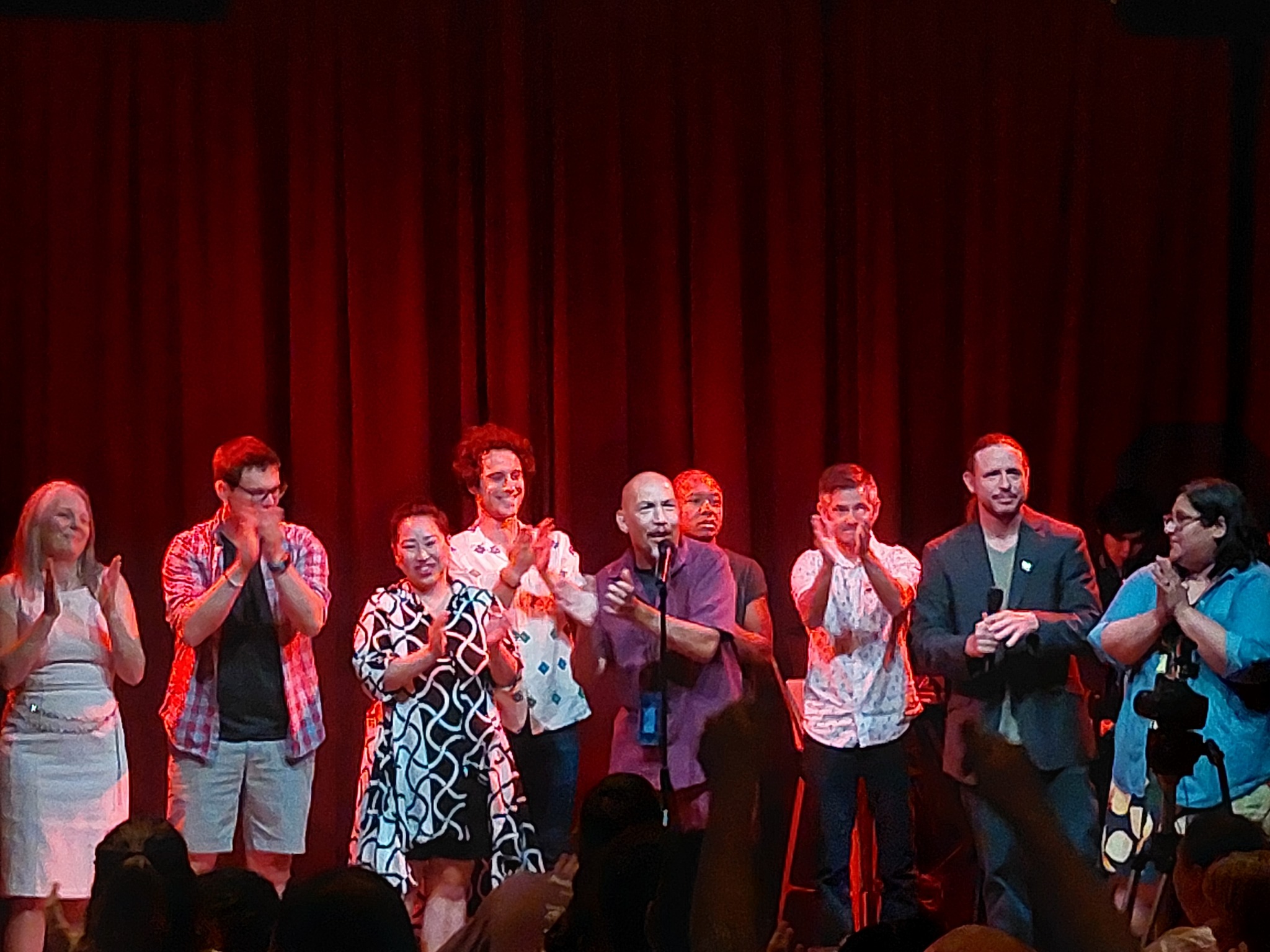 Group of people standing on stage with red curtains, some clapping and smiling, in a celebratory setting.