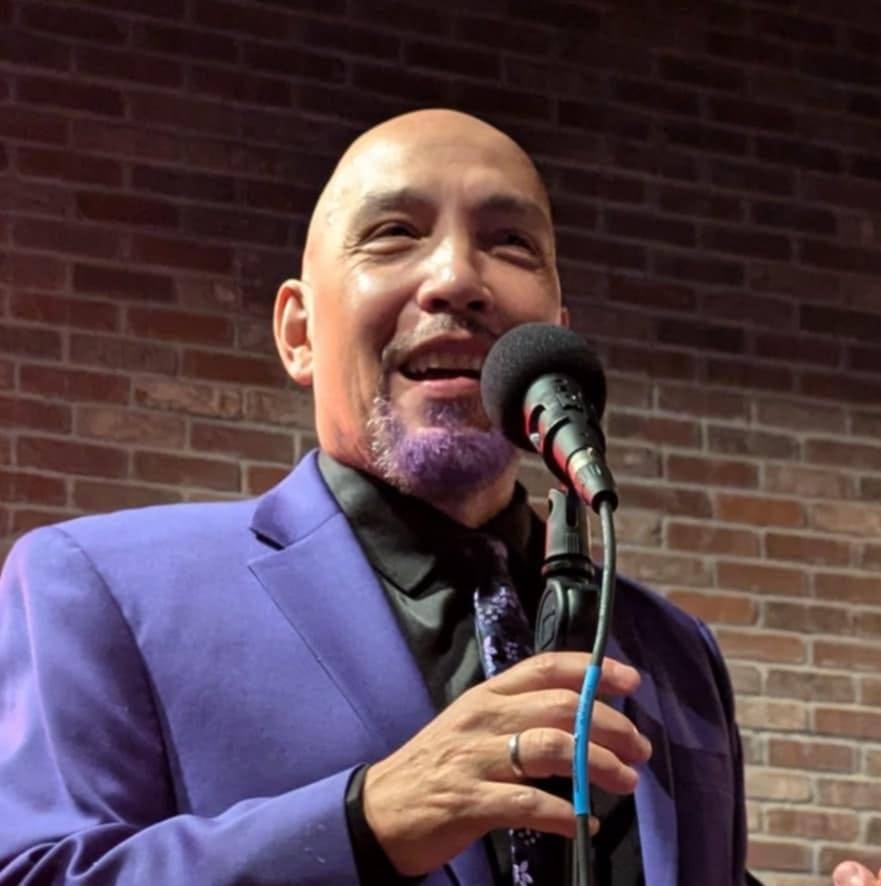Man with a shaved head and purple beard singing into a microphone, wearing a blue suit, against a brick wall background.