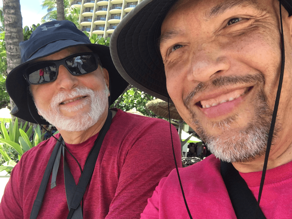Two men smiling outdoors, one wearing a black hat and sunglasses, the other wearing a black hat, both in red shirts.