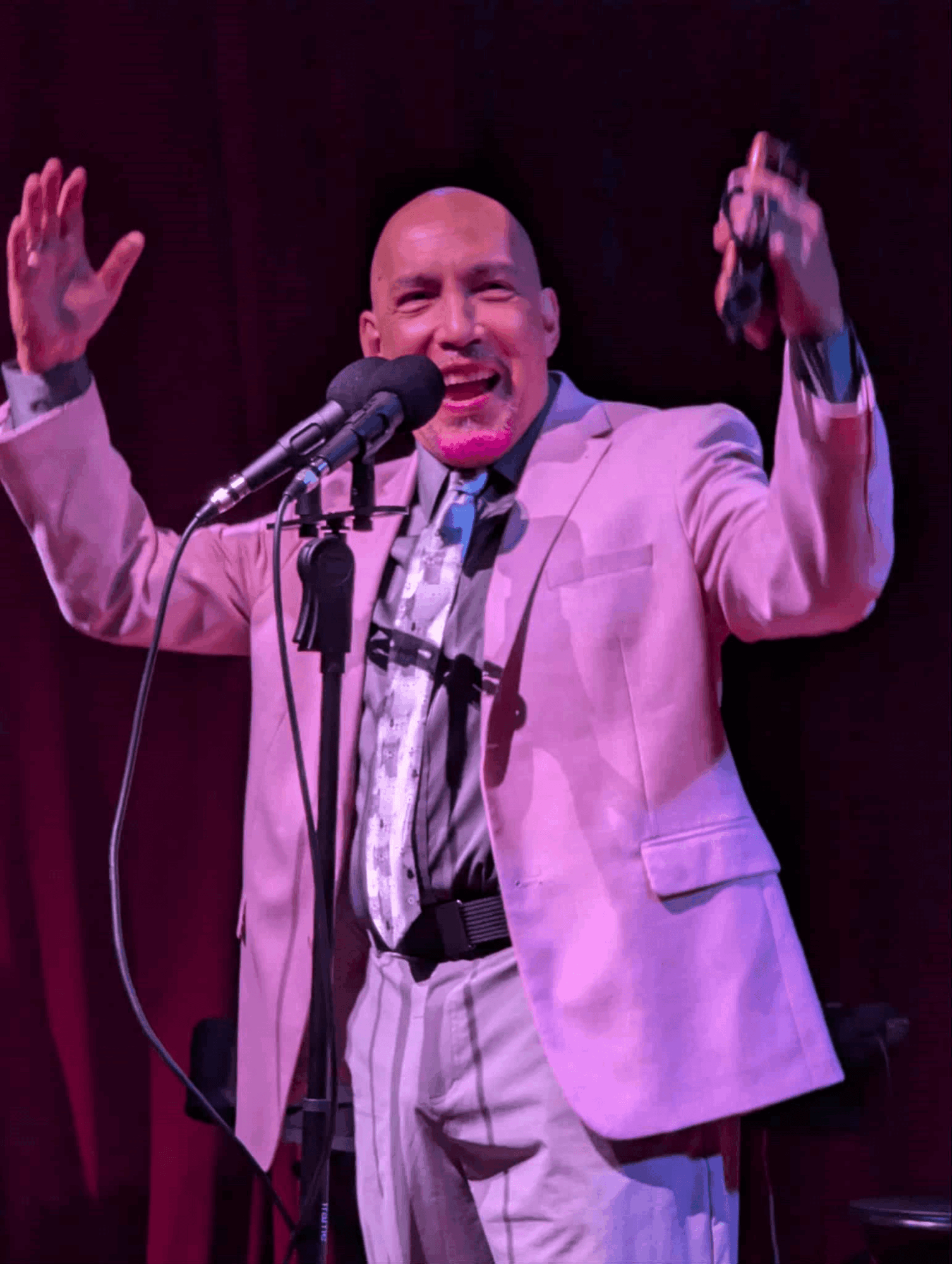 Person in a pink suit singing or speaking into a microphone with hands raised, on stage with dark background.
