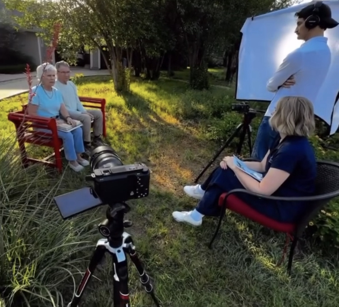Two people sit on a red bench outdoors, with a camera on a tripod and a person filming nearby, in a sunny park.