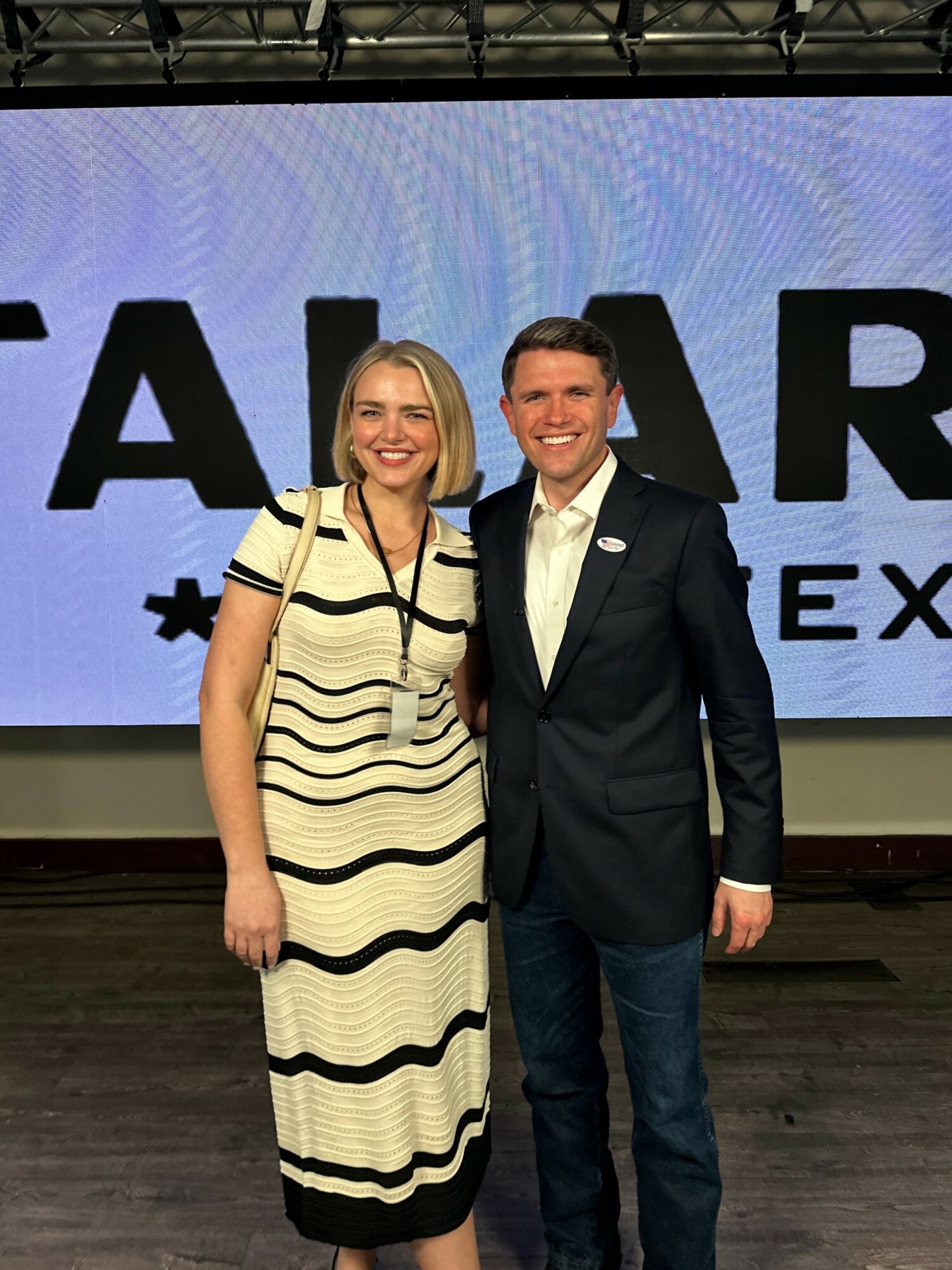 Two people, a woman in a striped dress and a man in a suit, stand together in front of a large screen.