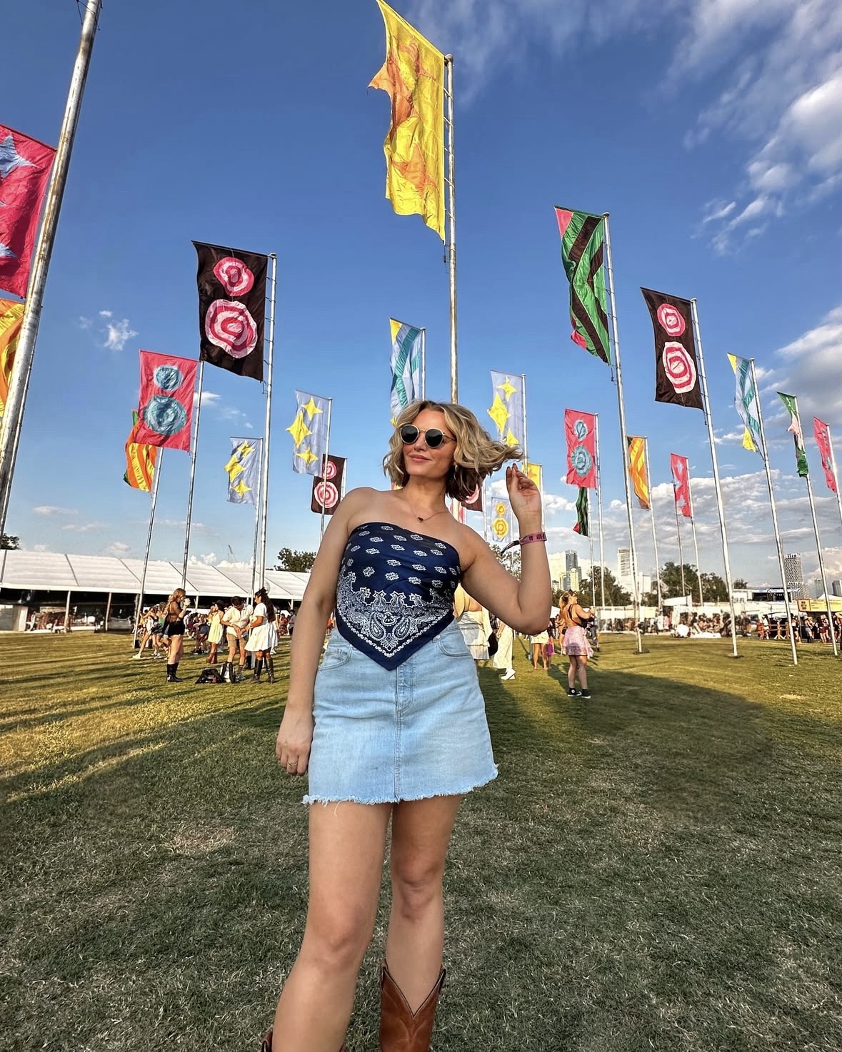 Woman in sunglasses and a blue dress standing outdoors with colorful flags and a crowd in the background.