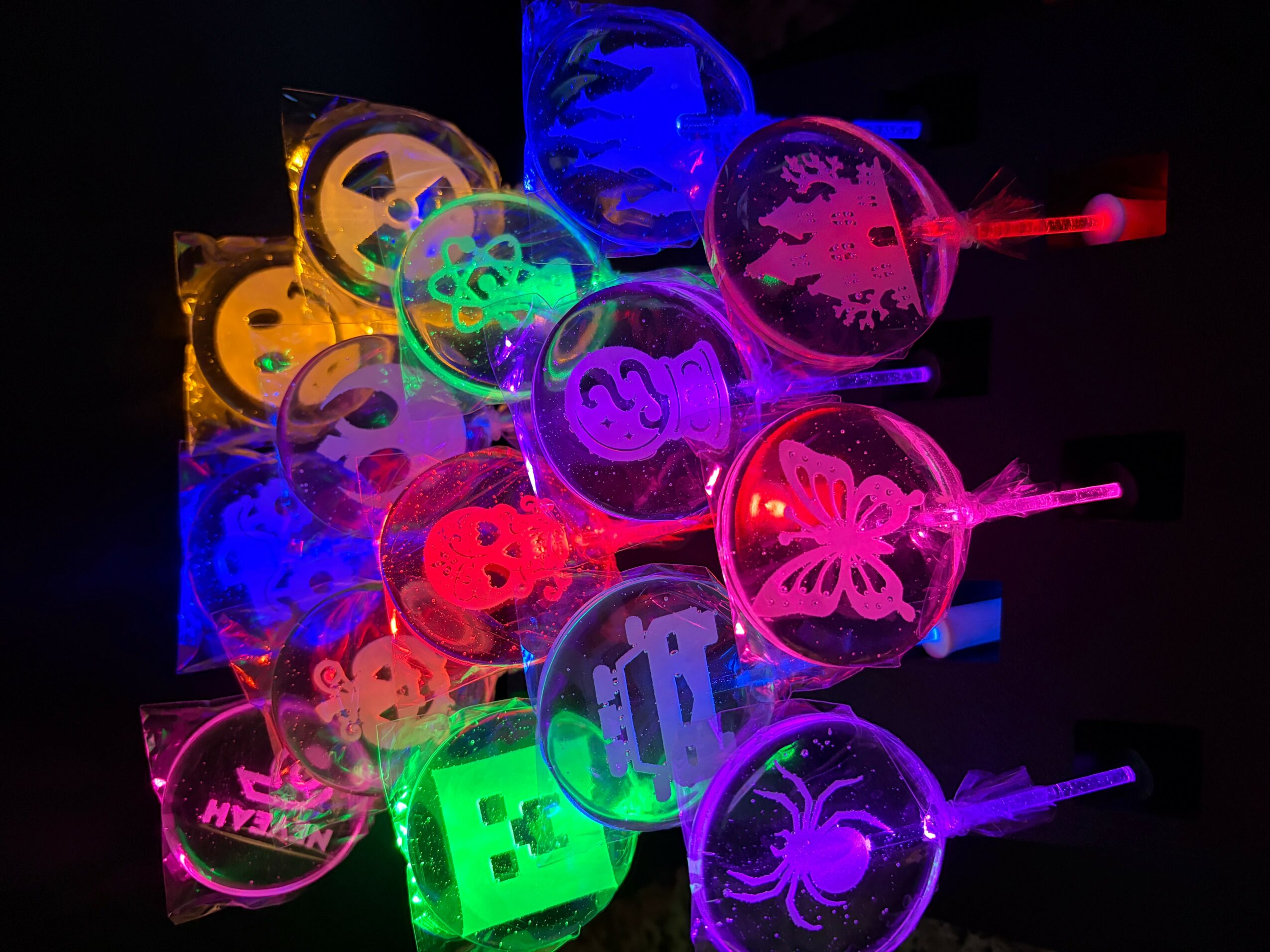 Multiple colorful glow-in-the-dark drink stirrers with various designs, arranged in a cluster against a dark background.