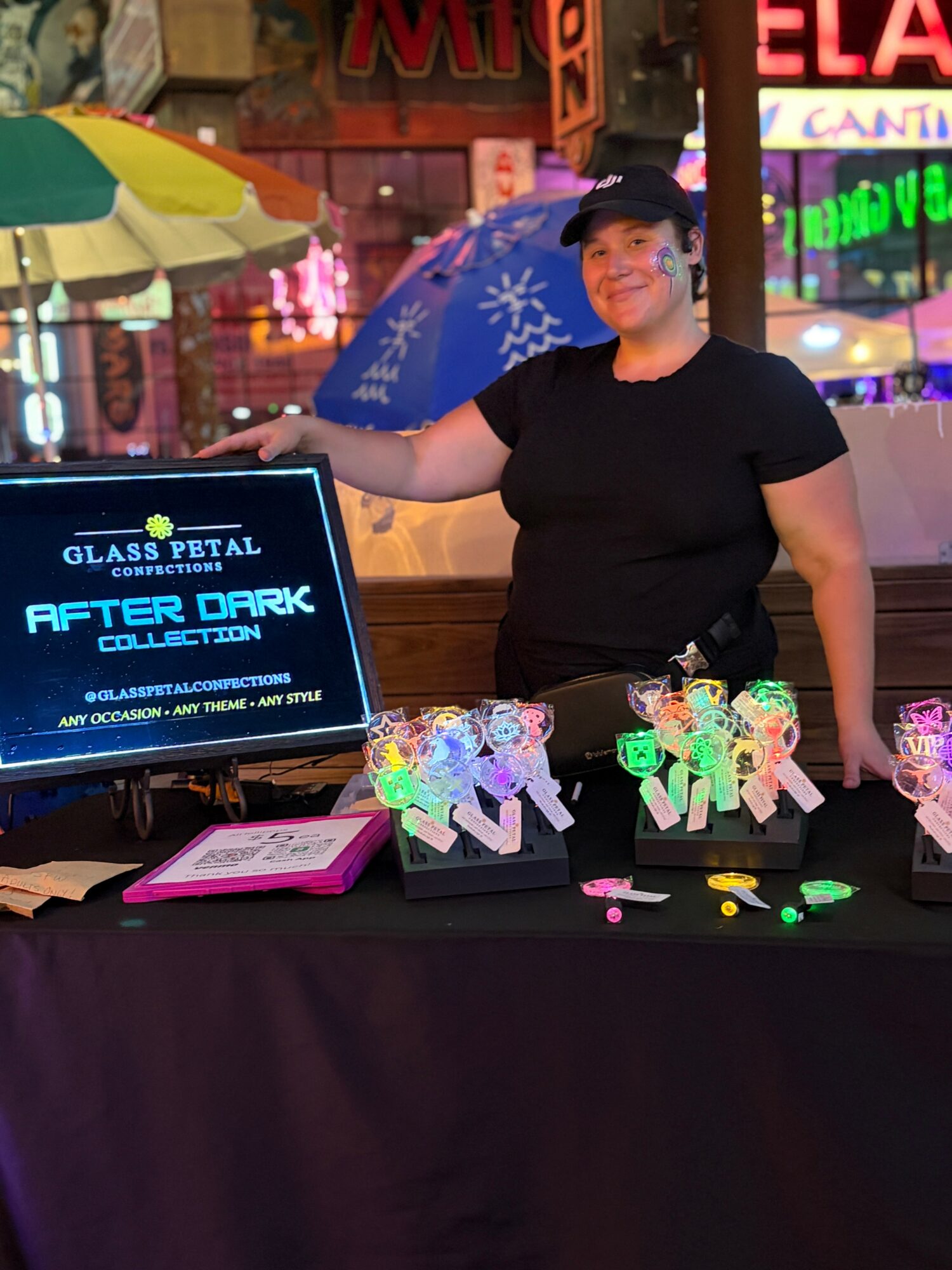 Person standing behind a table with colorful glass petal crafts, digital screen, and pink notebook, in an outdoor setting with umbrellas and neon signs.